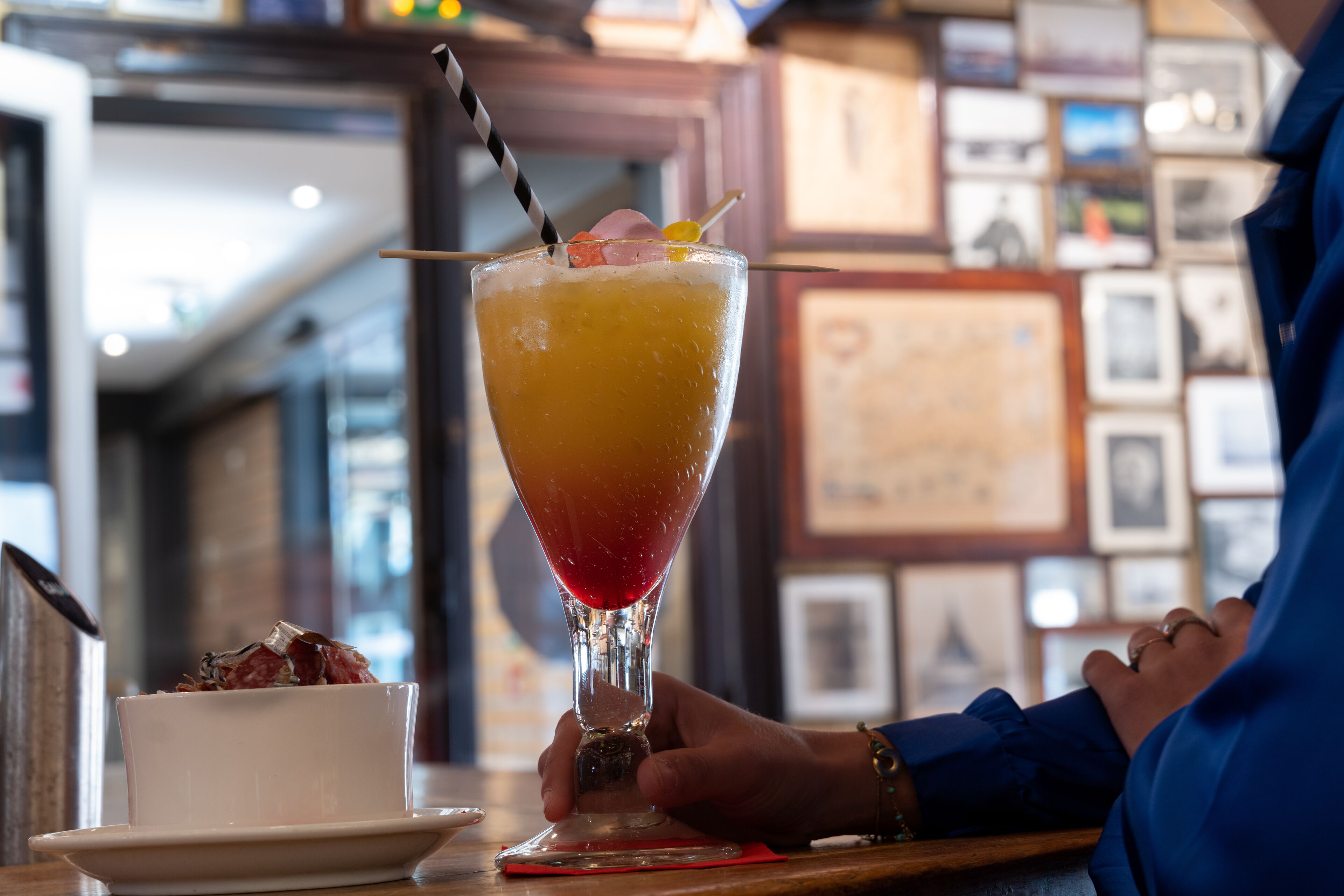 A cocktail in a glass with a straw, garnished with fruit, on a table with a framed picture in the background.
