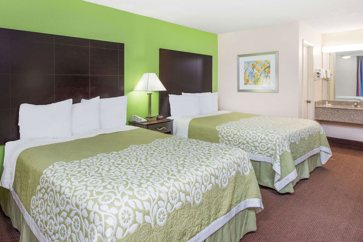 Habitación de hotel con dos camas, una mesita de noche con lámpara, un cuadro y un baño con espejo y lavamanos.