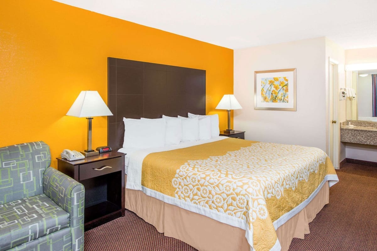 Habitación de hotel con una cama, dos mesitas de noche con lámparas, una silla, un cuadro en la pared y un baño visible a través de una puerta abierta.