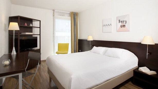 A hotel room with a bed, a desk with a lamp, a TV, a chair, and a nightstand with a lamp. - Residhome Appart Hotel Nancy Lorraine (Nancy)