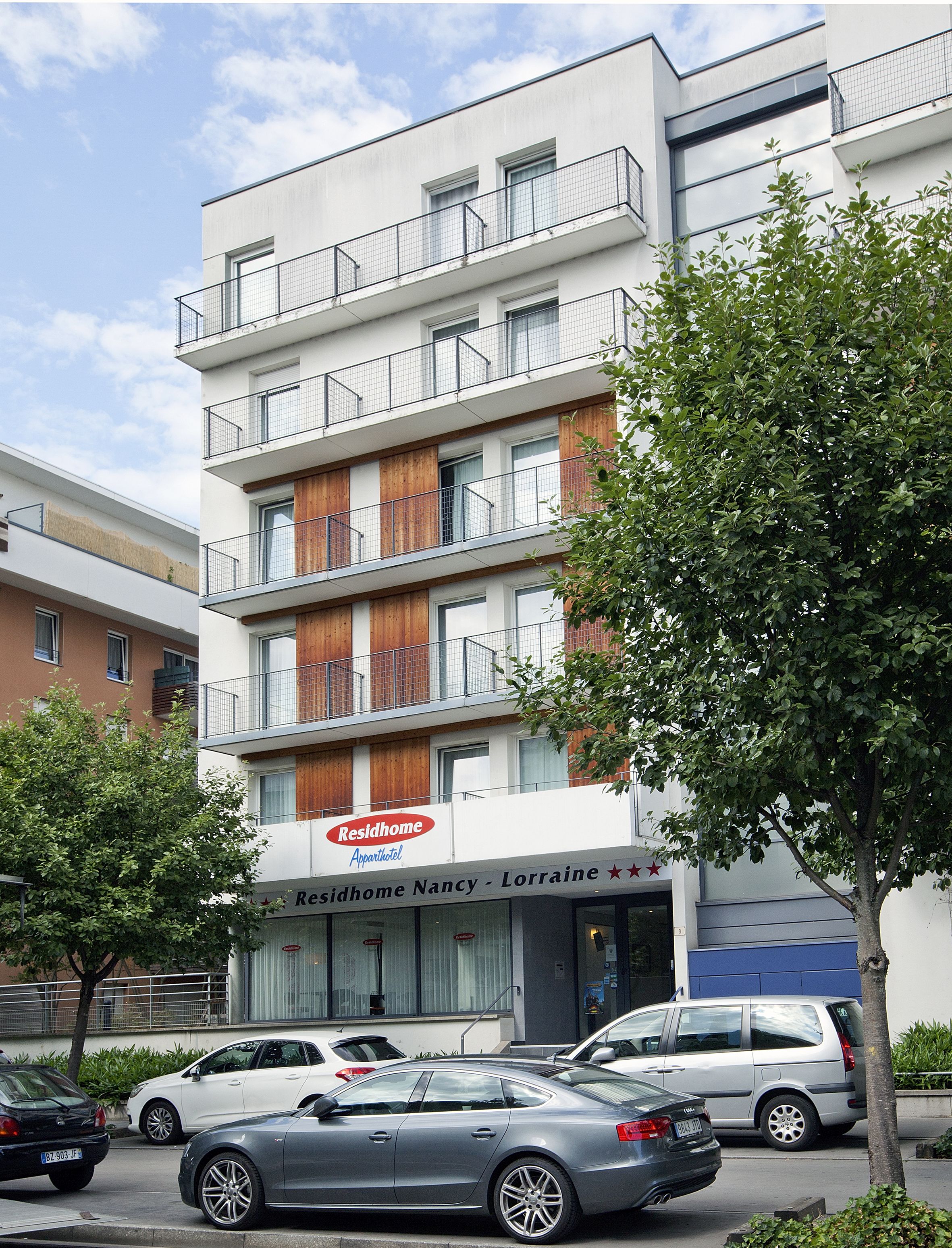 A multi-story building with a sign for "Residhome Nancy - Lorraine" indicating it is an apartment hotel. There are several cars parked in front, and a tree is visible to the right.