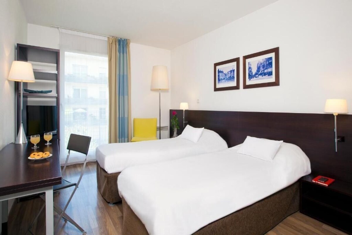 A hotel room with two beds, a desk with a lamp, a chair, and a window with curtains.