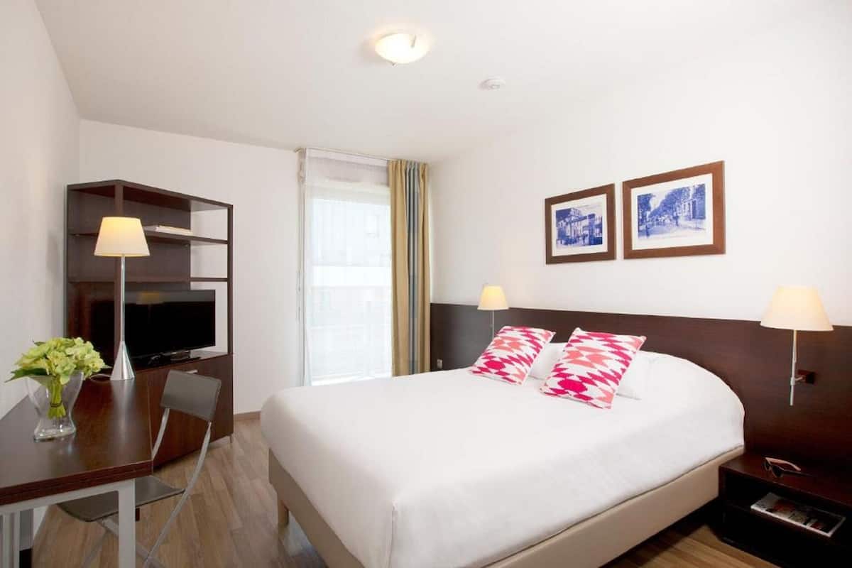 Een hotelkamer met een bed, een bureau met een televisie, een stoel, een lamp en twee ingelijste foto's aan de muur.