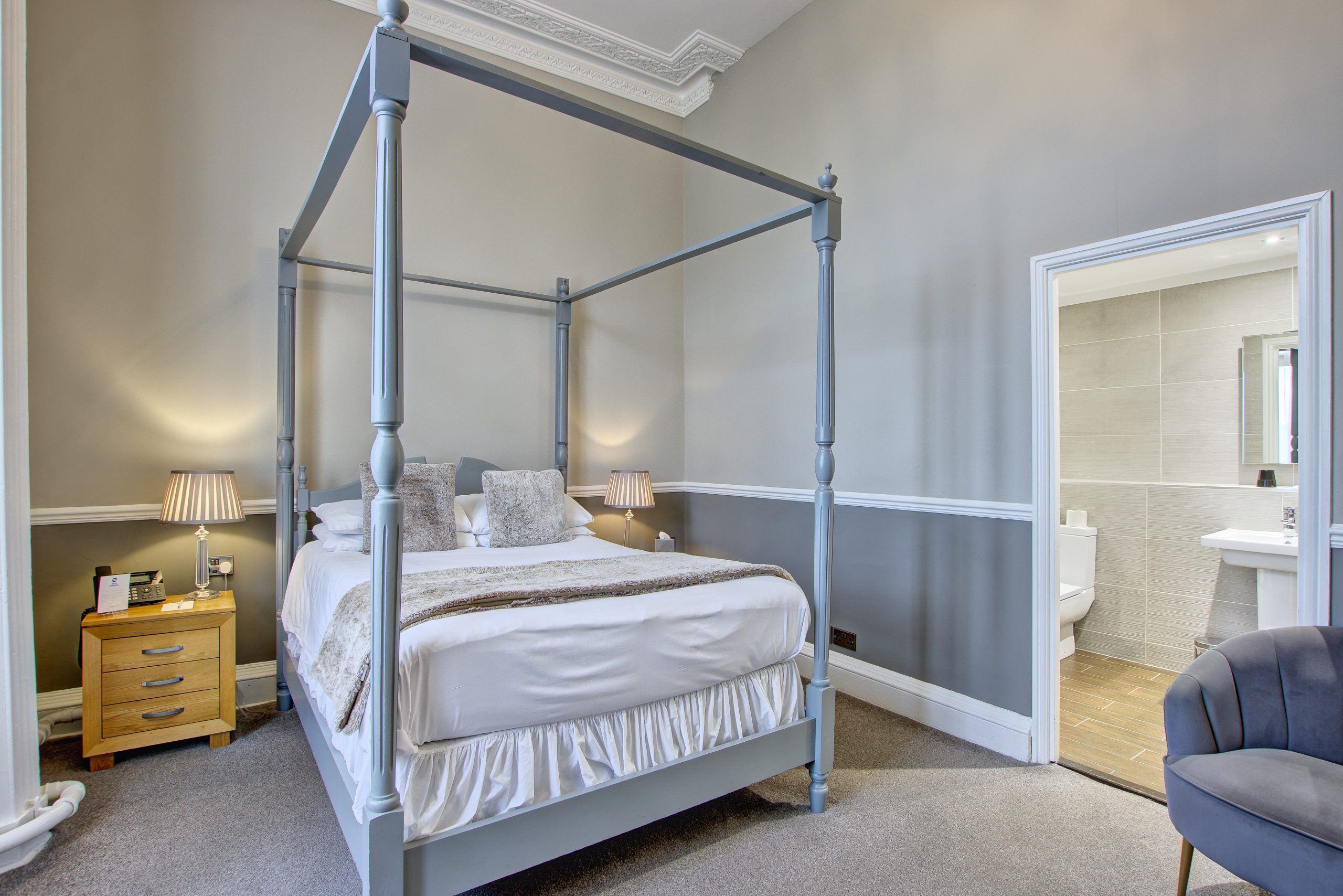 Een slaapkamer met een hemelbed met houten frame, een nachtkastje met lades, een lamp en een stoel. Een badkamer is zichtbaar door een geopende deur.
