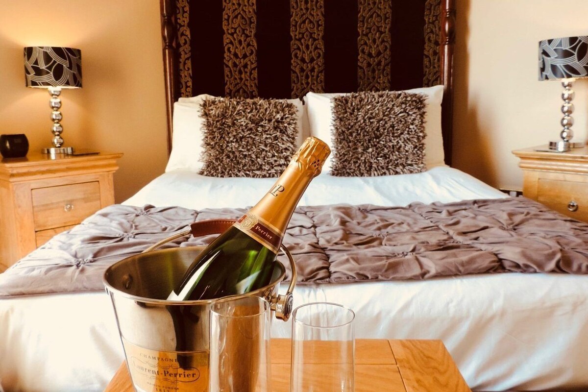 A hotel room with a bed, a bedside table with a lamp, and a bottle of champagne in a bucket.