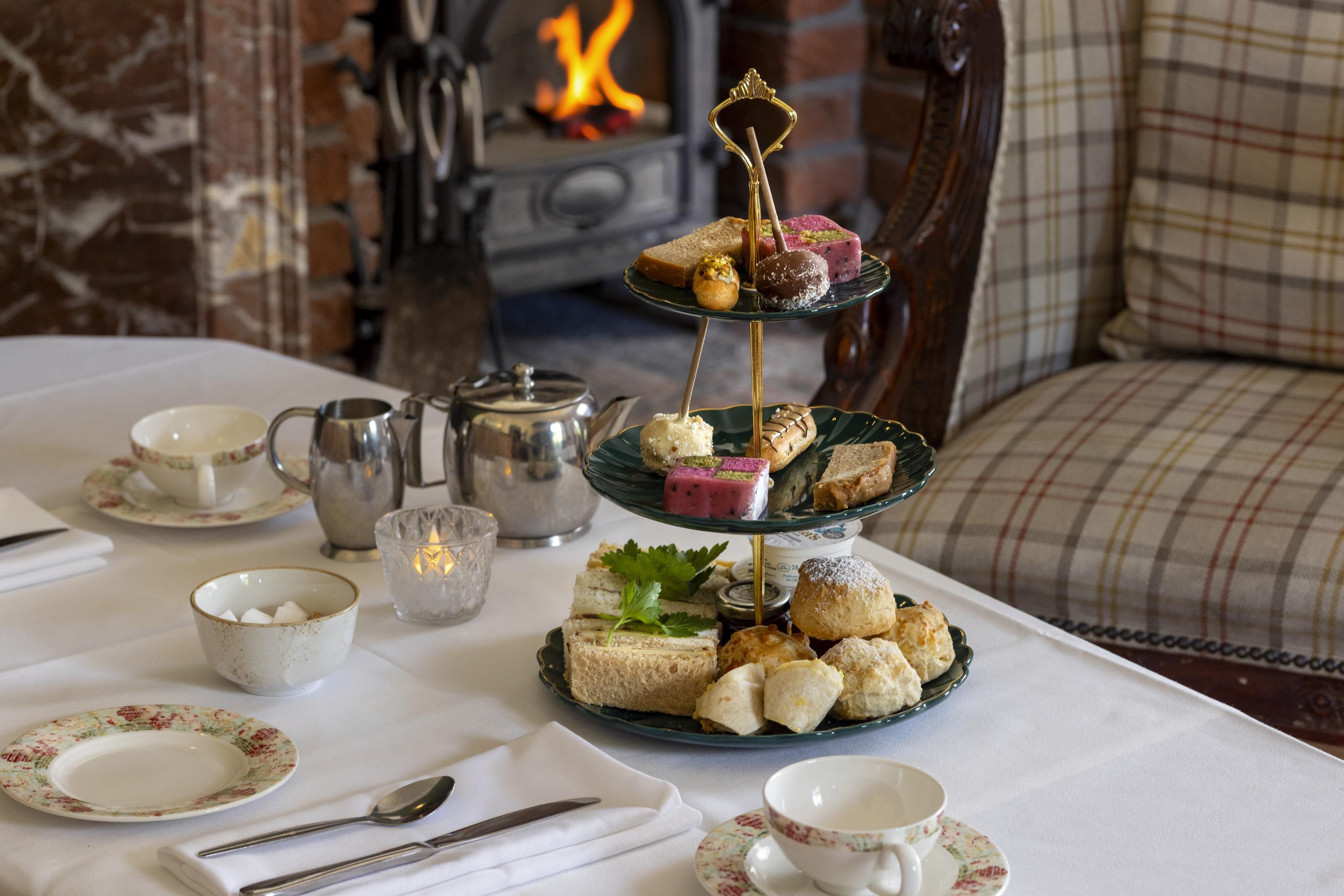 A three-tiered dessert stand with various pastries on a table, surrounded by tea cups, a teapot, and a candle.