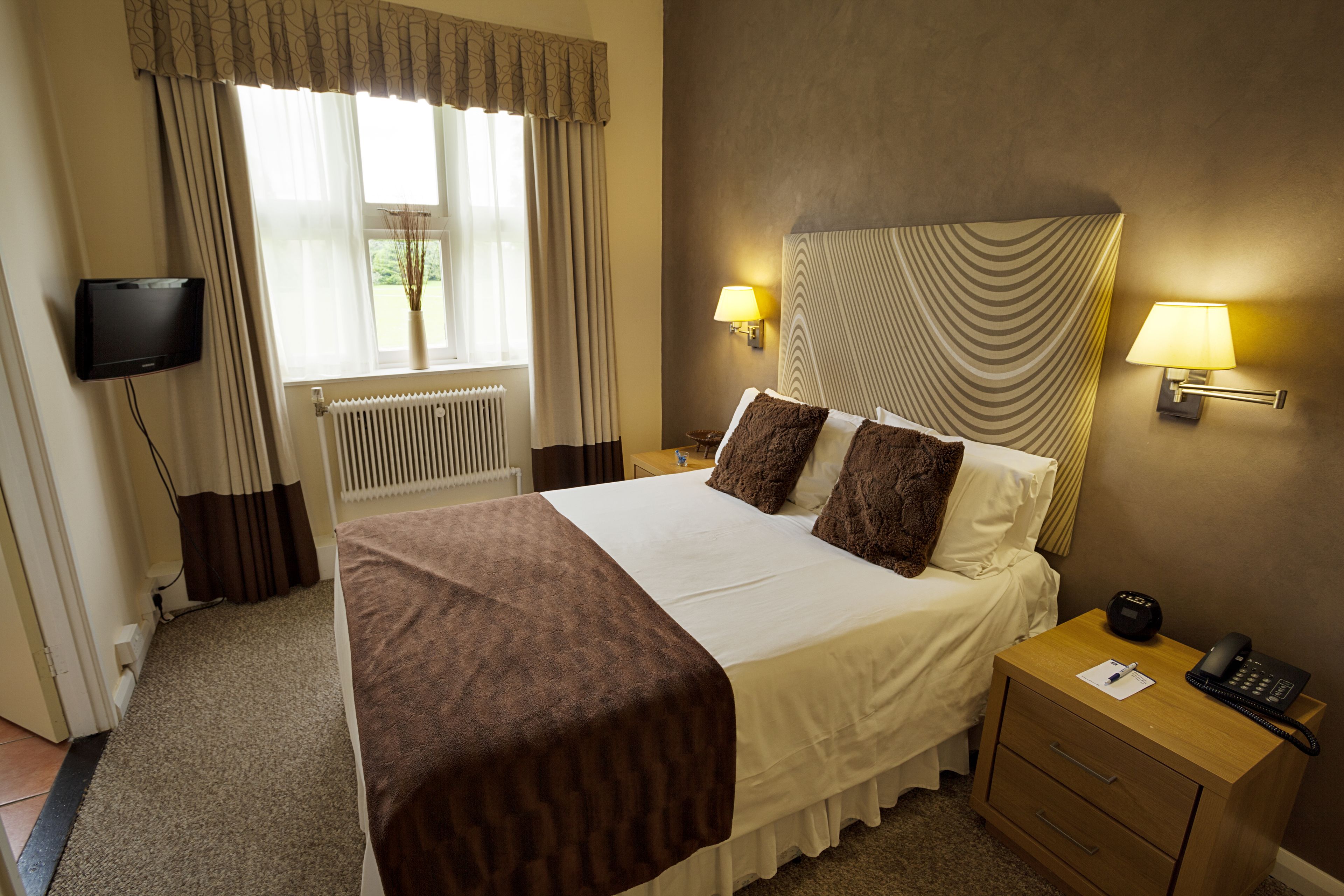 A hotel room with a bed, bedside lamps, a TV, a bedside table with a phone, and a window with curtains.