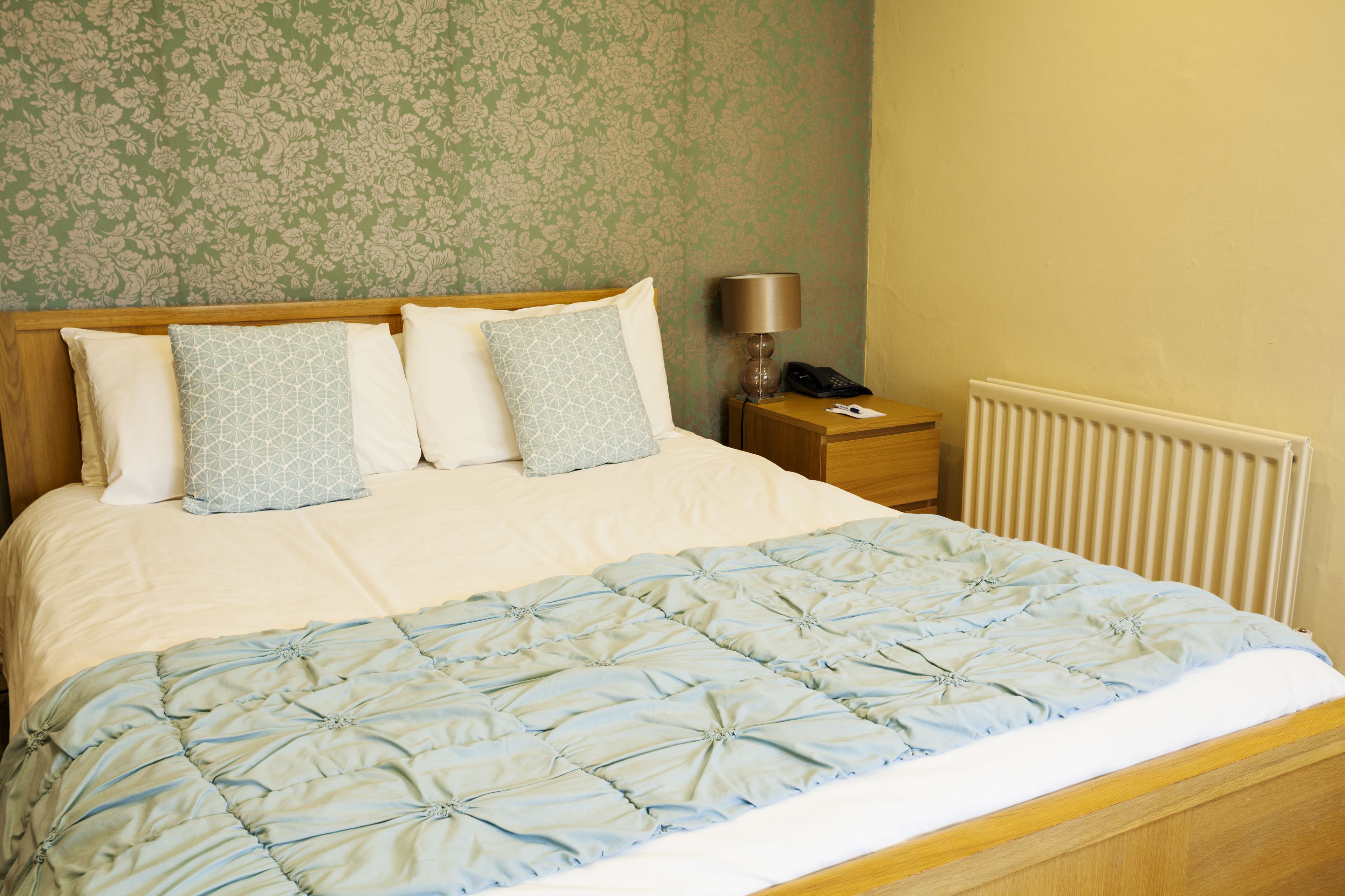 Una cama bien tendida con un edredón azul y dos almohadas, una lámpara de noche y una mesita de madera.
