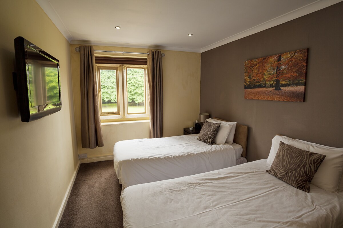 A hotel room with two beds, a flat-screen TV mounted on the wall, a window with curtains, and a painting on the wall.