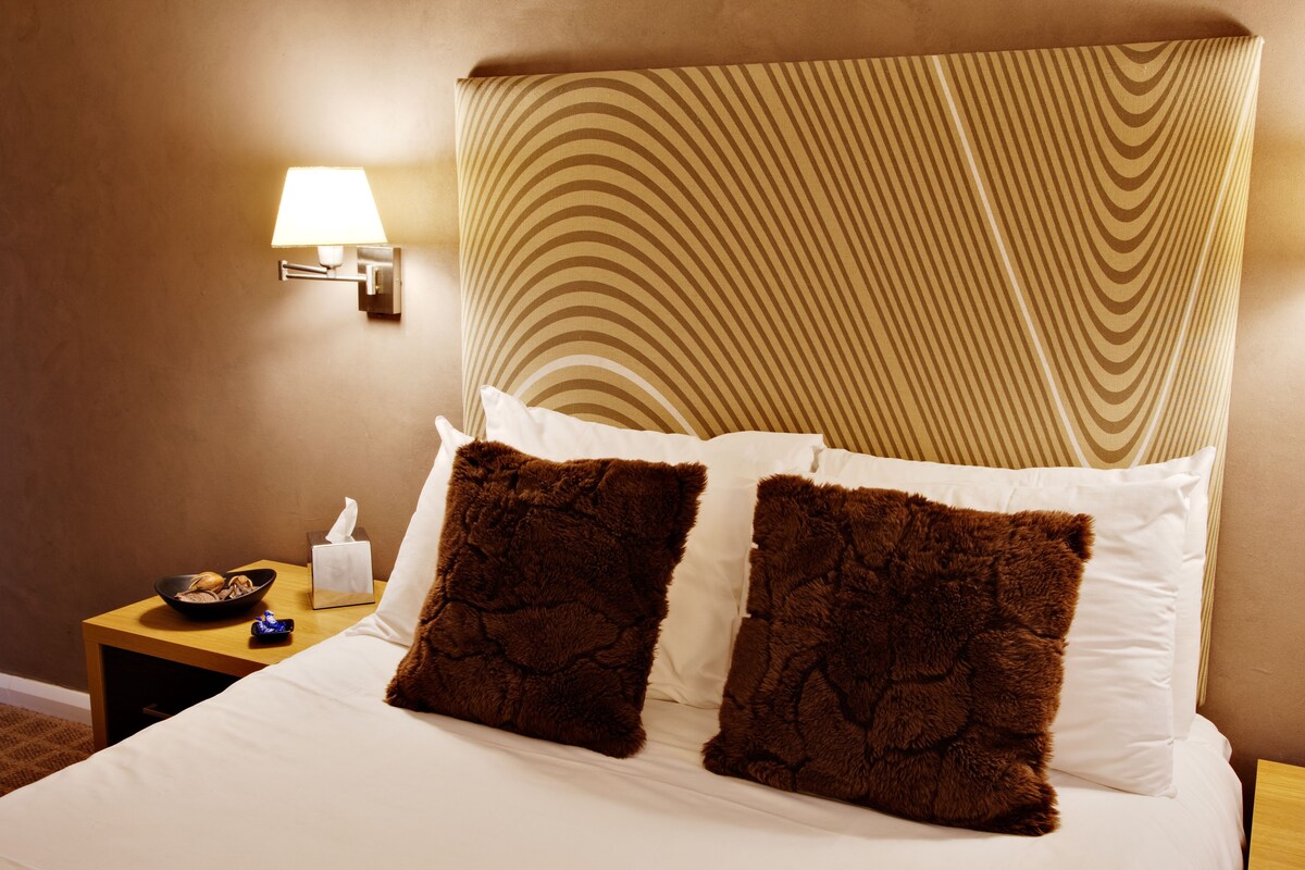 A neatly made bed with white and brown pillows, a bedside table with a bowl of fruit and a tissue box, and a wall-mounted lamp.
