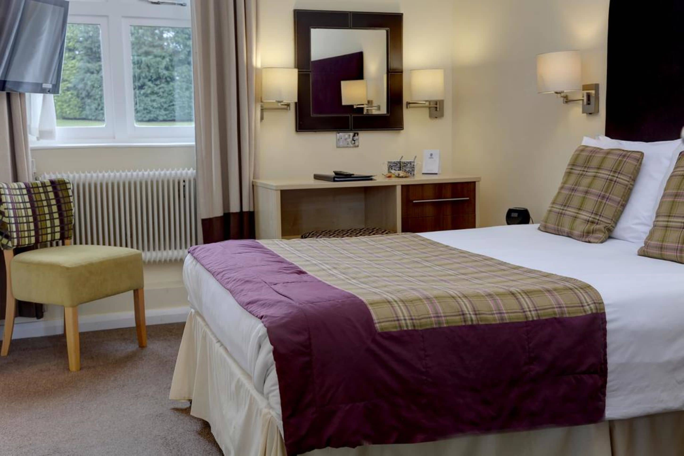 Een hotelkamer met een bed, nachtkastje, stoel, bureau, spiegel en raam.