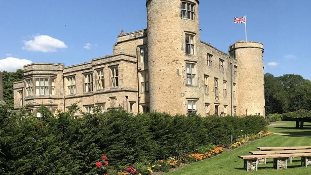 A historic stone building with a central tower and flag, surrounded by well-maintained gardens and a picnic area.