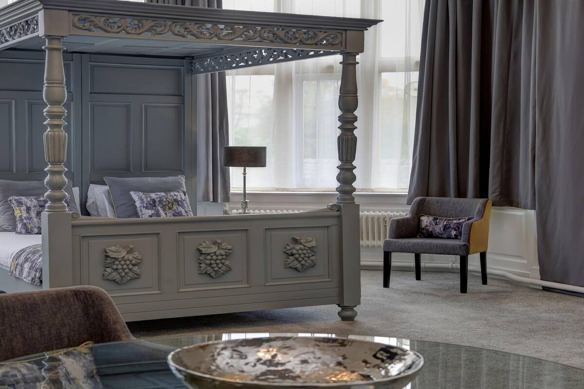 Una cama con dosel de acabado gris y detalles decorativos de hojas, un sillón gris y una mesa de centro de vidrio con base metálica.