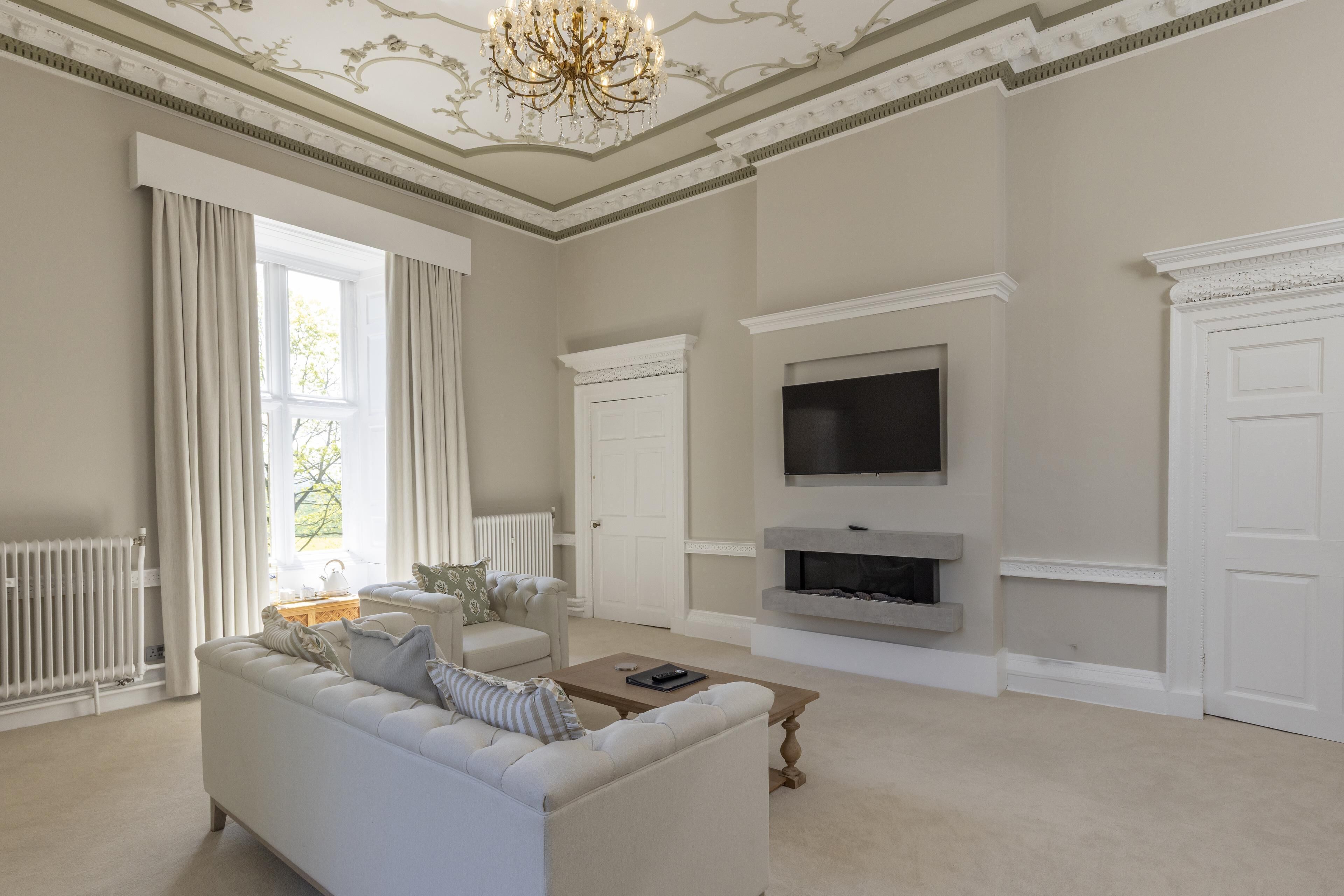 A spacious living room with a white sofa, a flat-screen TV mounted above a fireplace, and a large window with curtains.