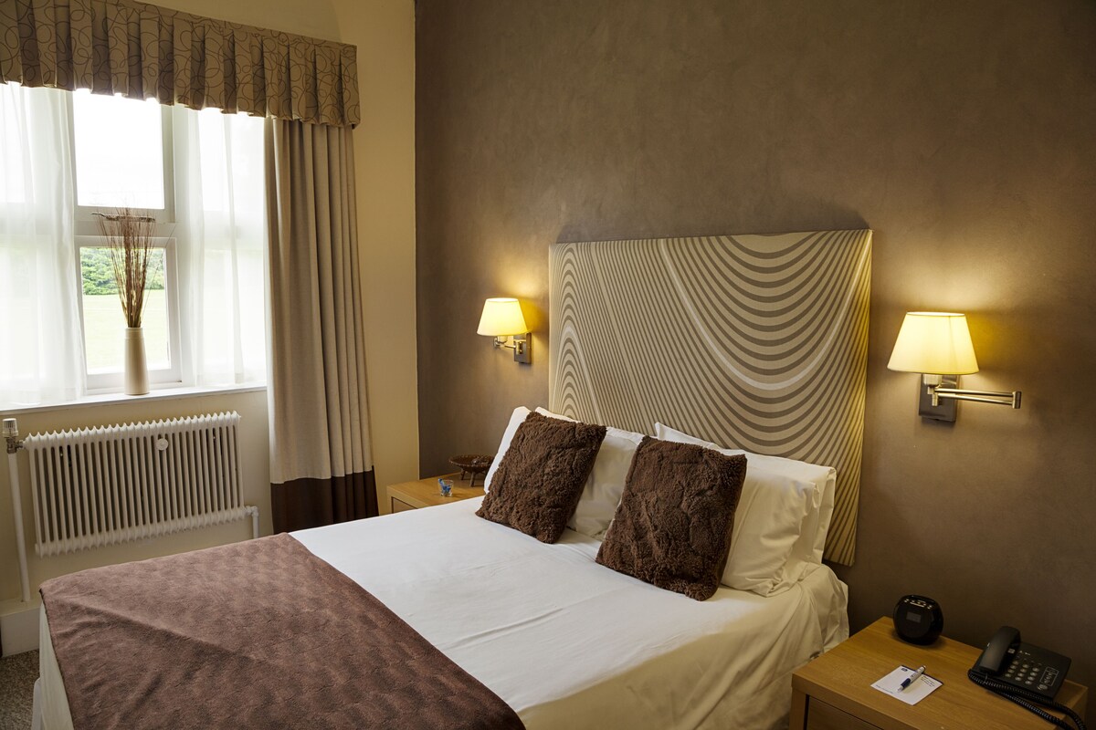 A hotel room with a bed, two bedside lamps, a nightstand with a phone and a notepad, a window with curtains, and a radiator.