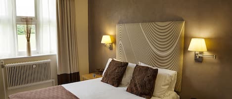A hotel room with a bed, two bedside lamps, a nightstand with a phone and a notepad, a window with curtains, and a radiator.