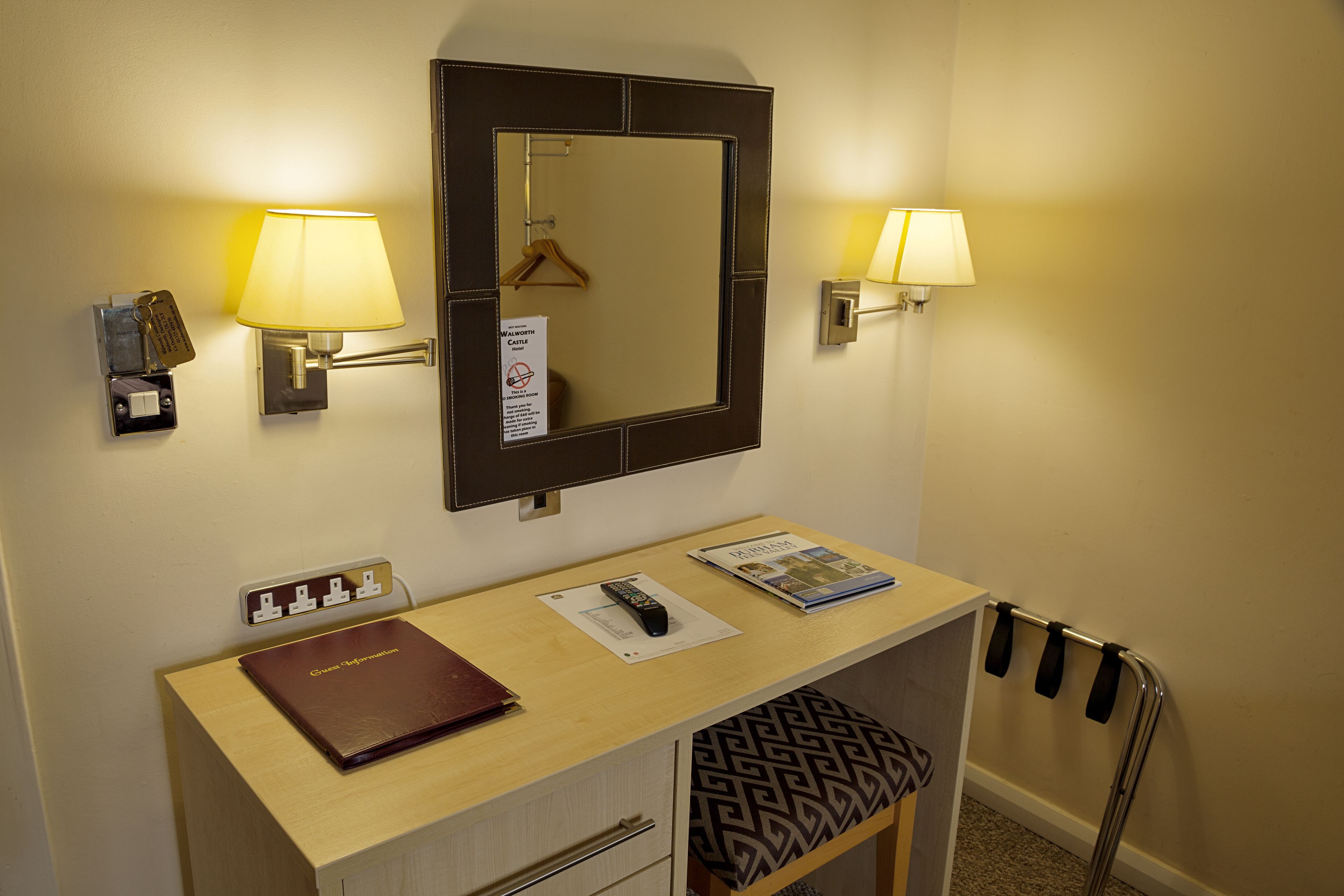 Habitación de hotel con escritorio, espejo, dos lámparas de pared, perchero, atril para abrigos, control remoto, revista y portal tarjetas.