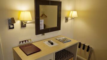 A hotel room with a desk, a mirror, two wall-mounted lamps, a hanging coat rack, a coat stand, a remote control, a magazine, and a keycard holder.