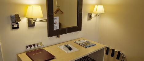 A hotel room with a desk, a mirror, two wall-mounted lamps, a hanging coat rack, a coat stand, a remote control, a magazine, and a keycard holder.