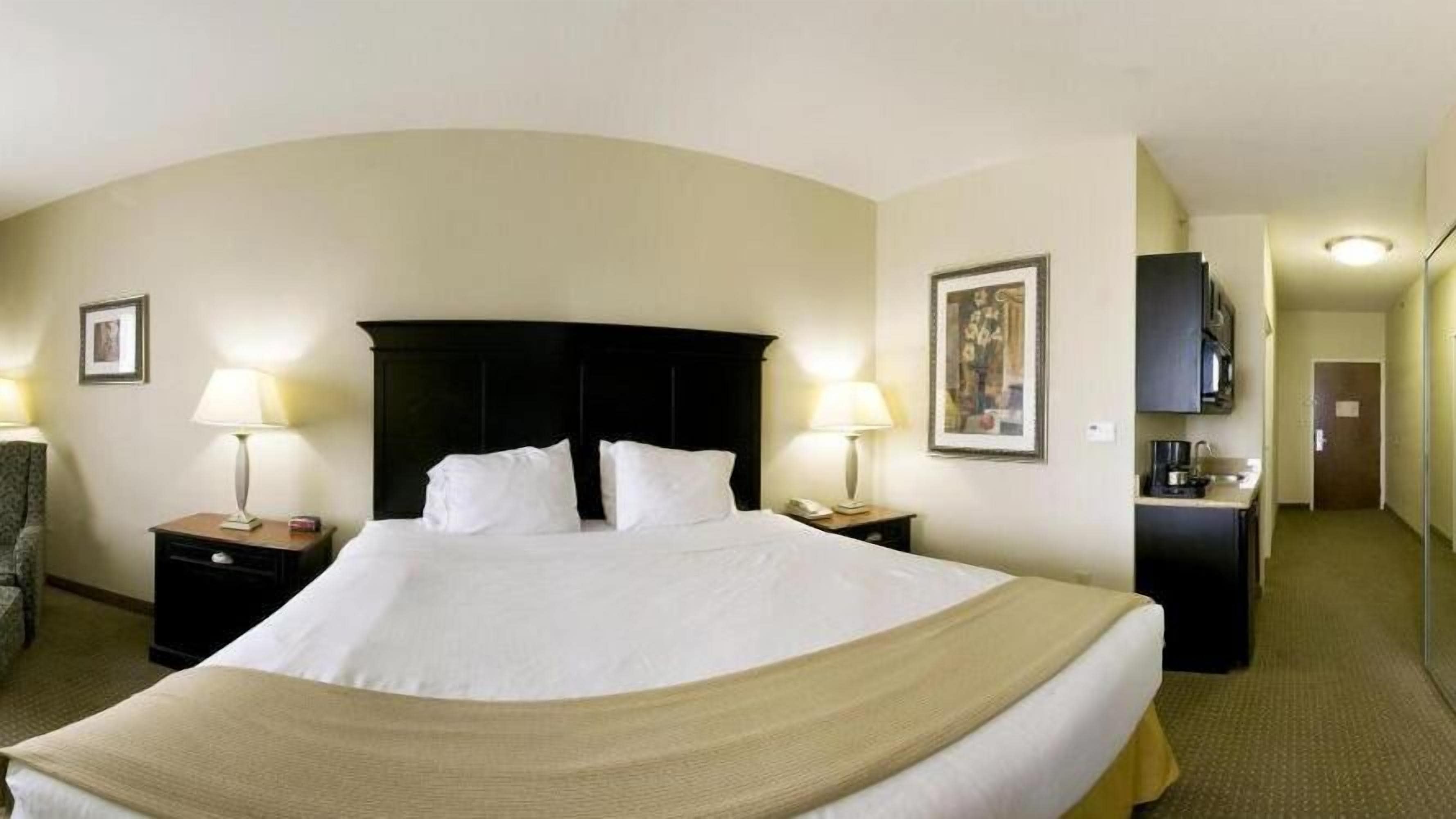 Une chambre d’hôtel avec un grand lit, deux tables de chevet avec des lampes, une chaise, un bureau avec un téléphone et un tableau encadré au mur.