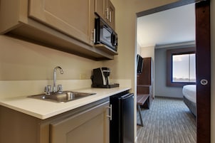 A compact hotel room with a kitchenette including a sink, microwave, and coffee maker. There is a bed with a window, and the room has a carpeted floor.