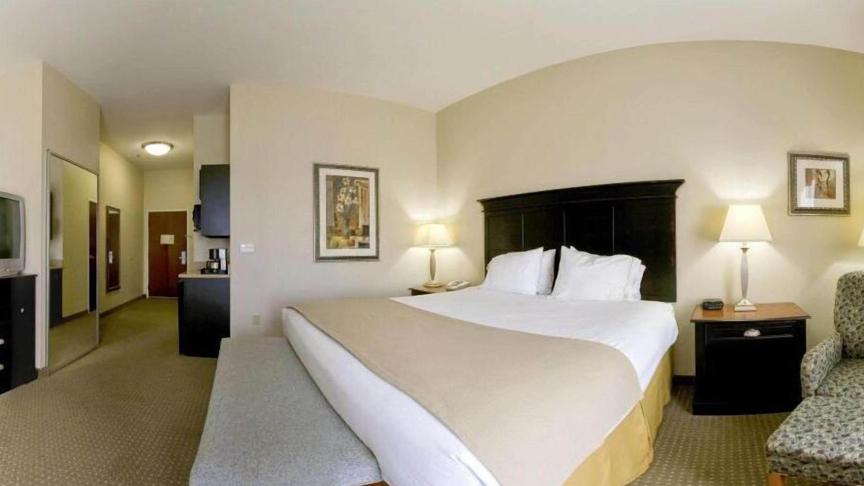Una habitación de hotel con una cama grande, dos mesitas de noche con lámparas, una silla, un escritorio con computadora y un televisor.