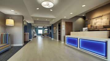 Un área de recepción de hotel con diseño moderno, que incluye un mostrador de recepción con paneles azules iluminados, un área de descanso con un sofá a rayas y un pasillo que conduce a un comedor.
