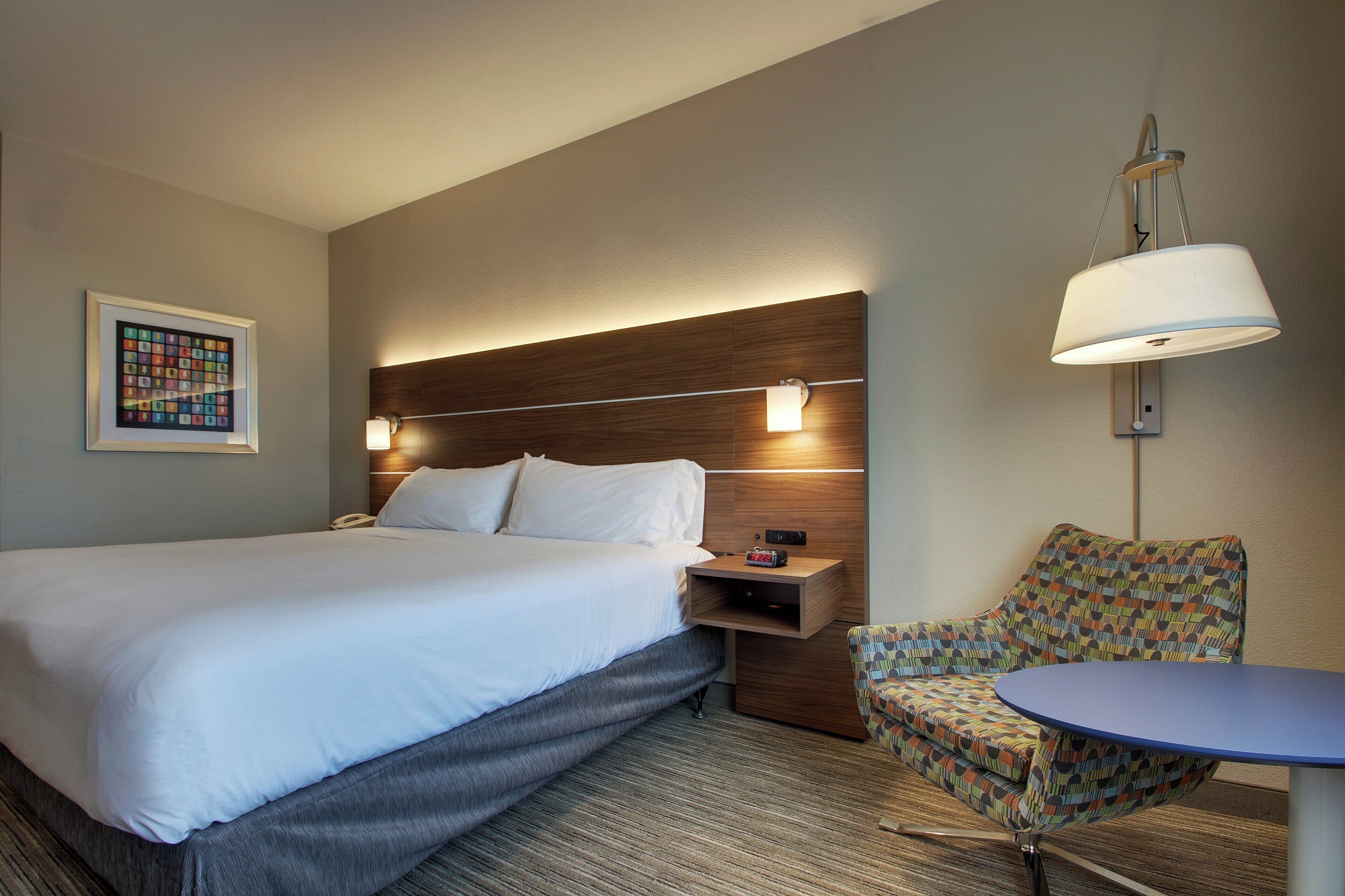 Habitación de hotel con una cama grande, mesita de noche, lámpara, silla y una pequeña mesa.