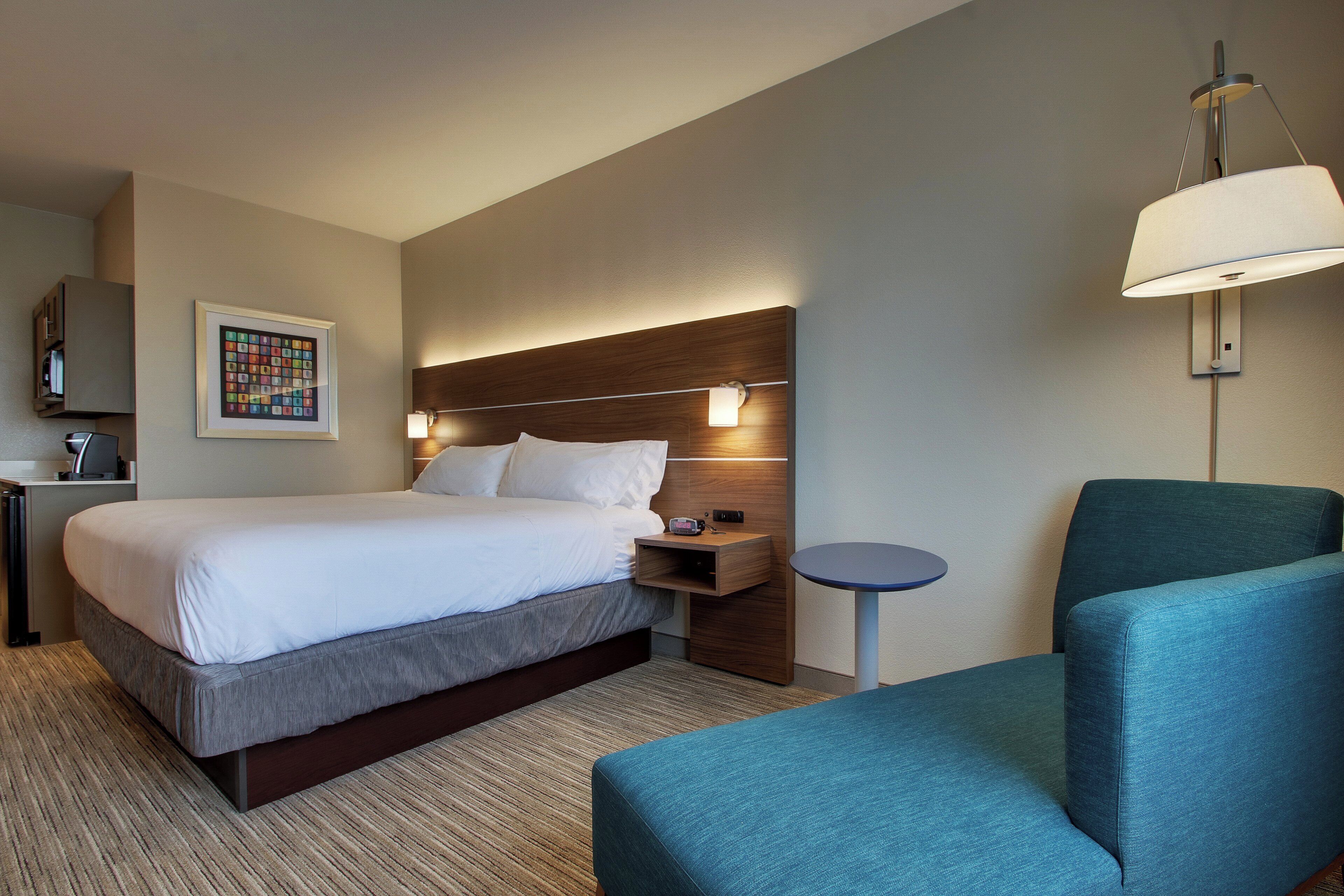 A hotel room with a bed, bedside table, lamp, chair, and a wall-mounted artwork.