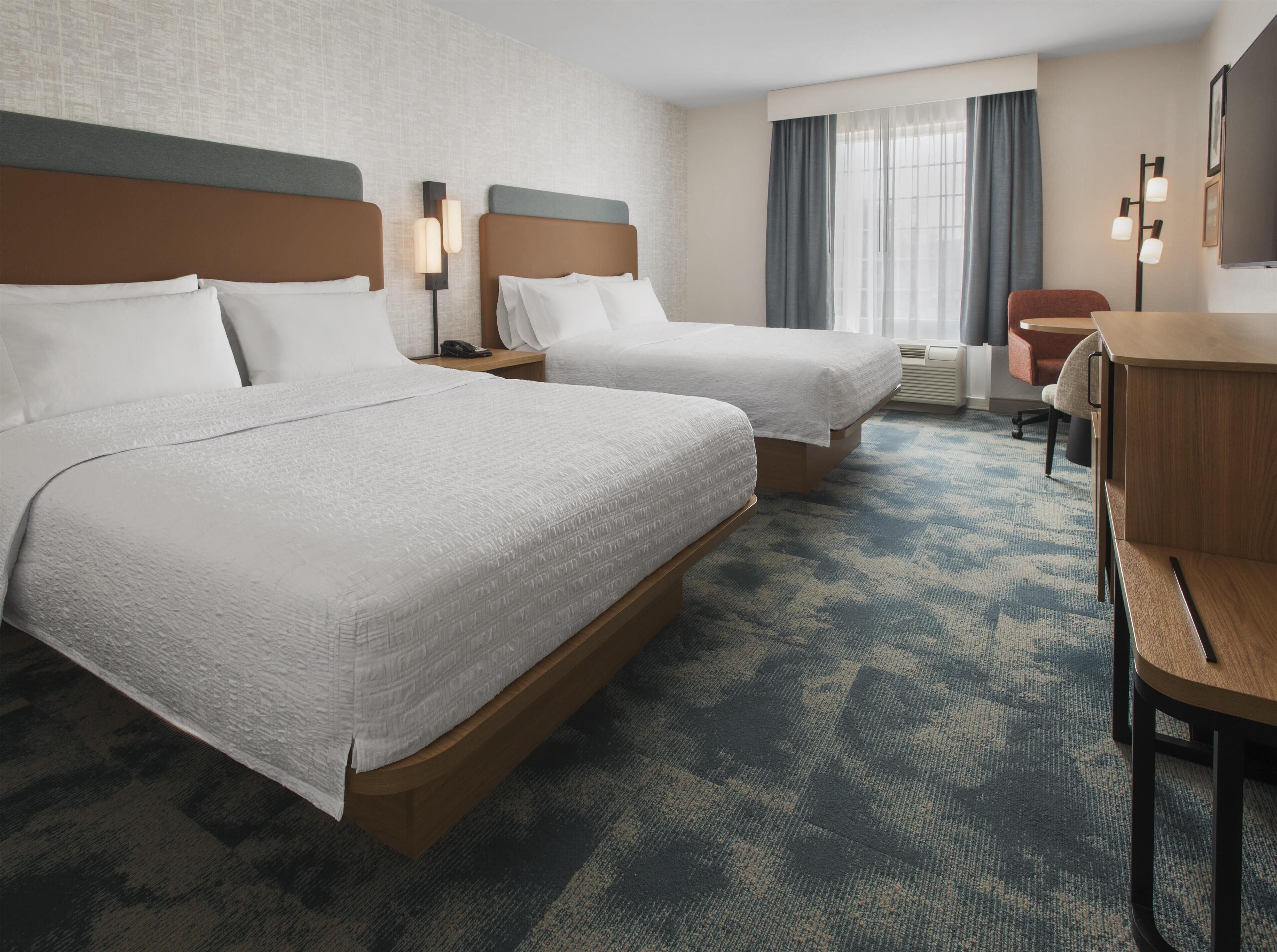 A hotel room with two beds, a desk, a chair, and a large window with curtains.