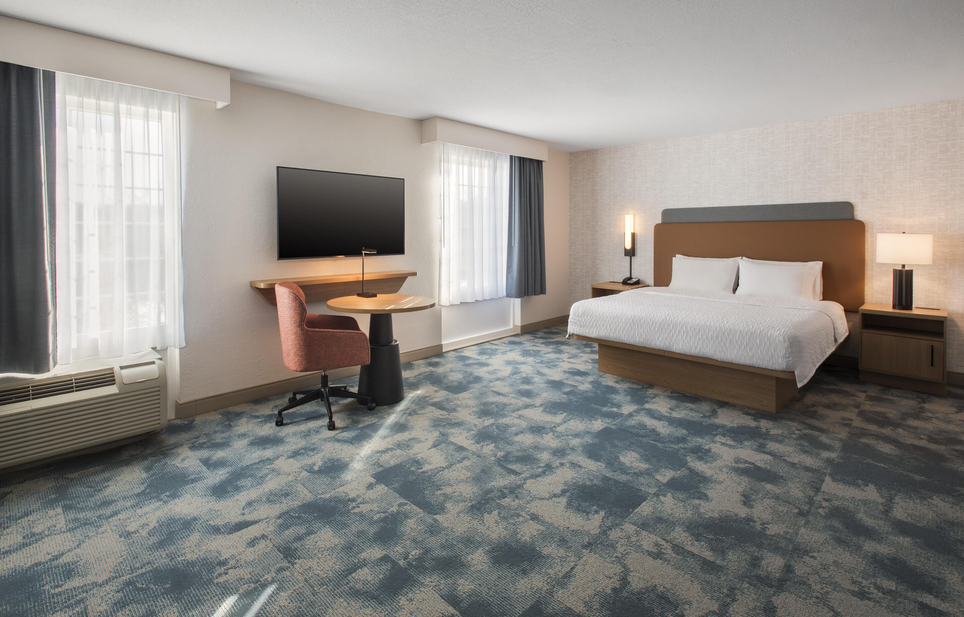 A hotel room with a bed, a TV, a desk, a chair, and a window with curtains.