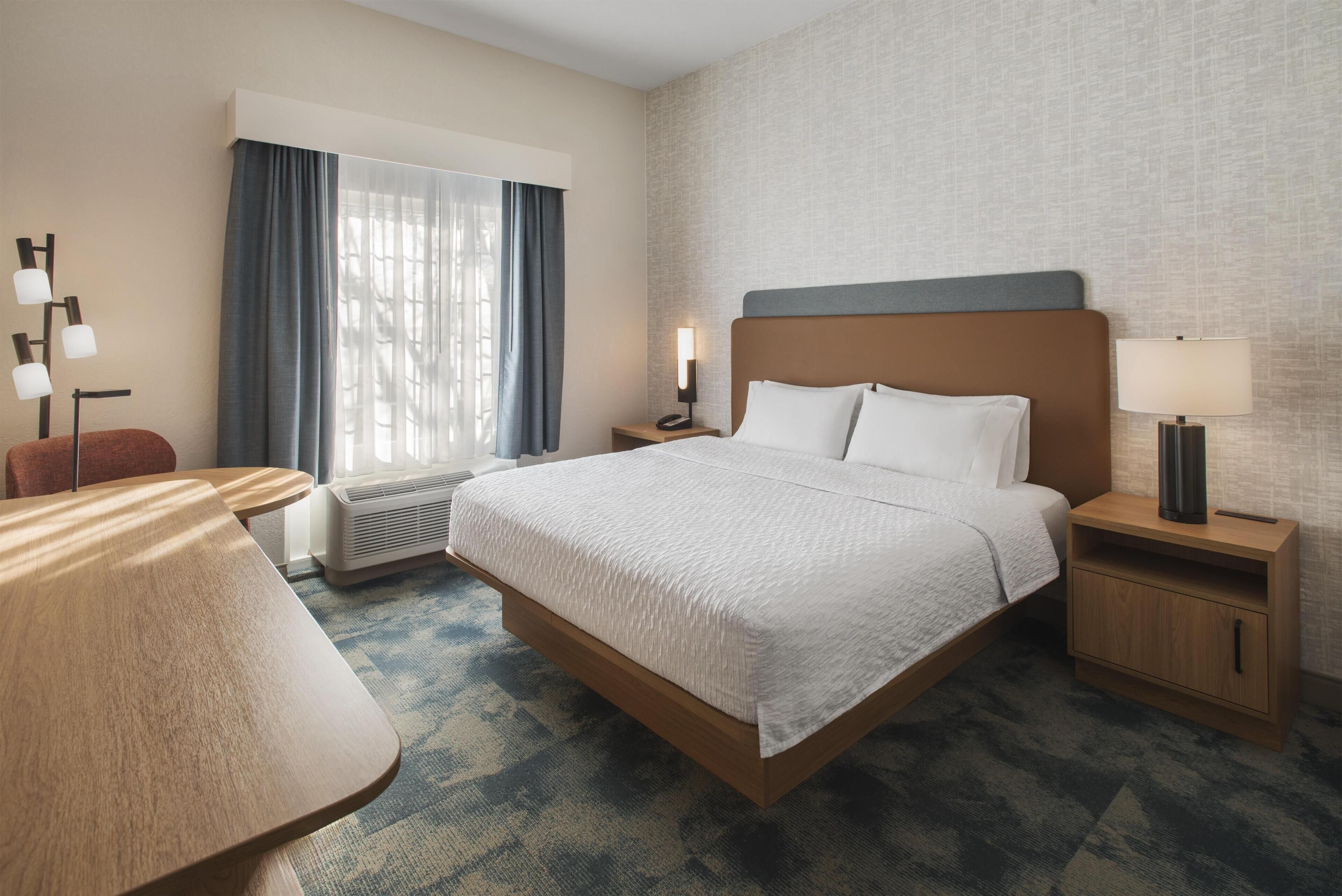Habitación de hotel con cama, escritorio, silla, mesita de noche con lámpara y calefactor en la pared.