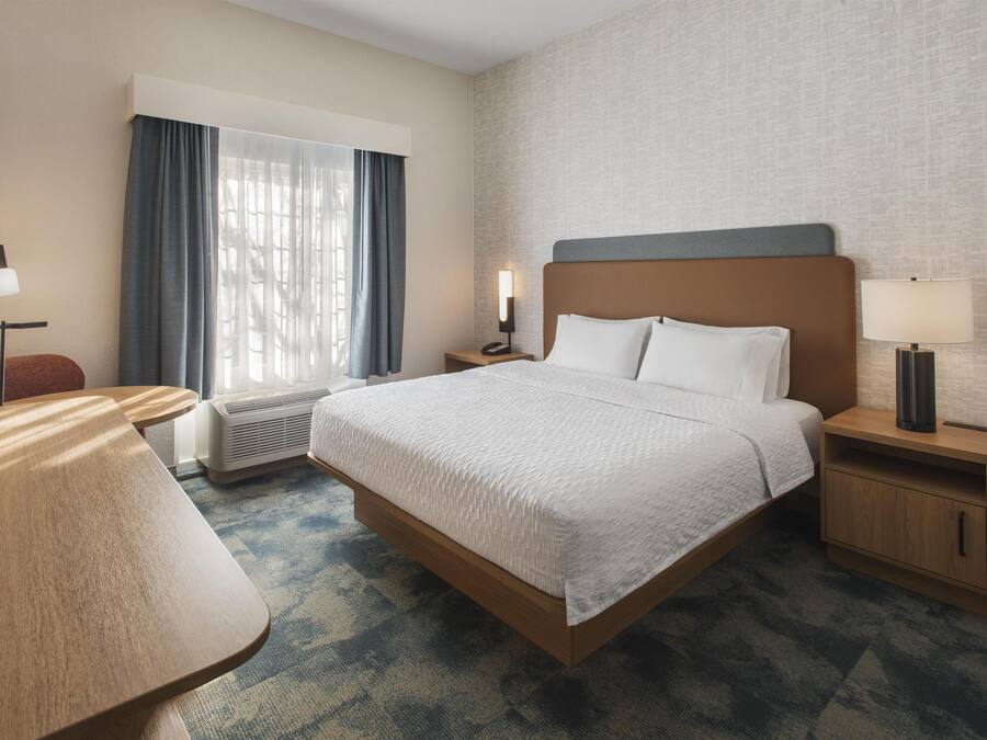Habitación de hotel con cama, escritorio, silla, mesita de noche con lámpara y calefactor en la pared.