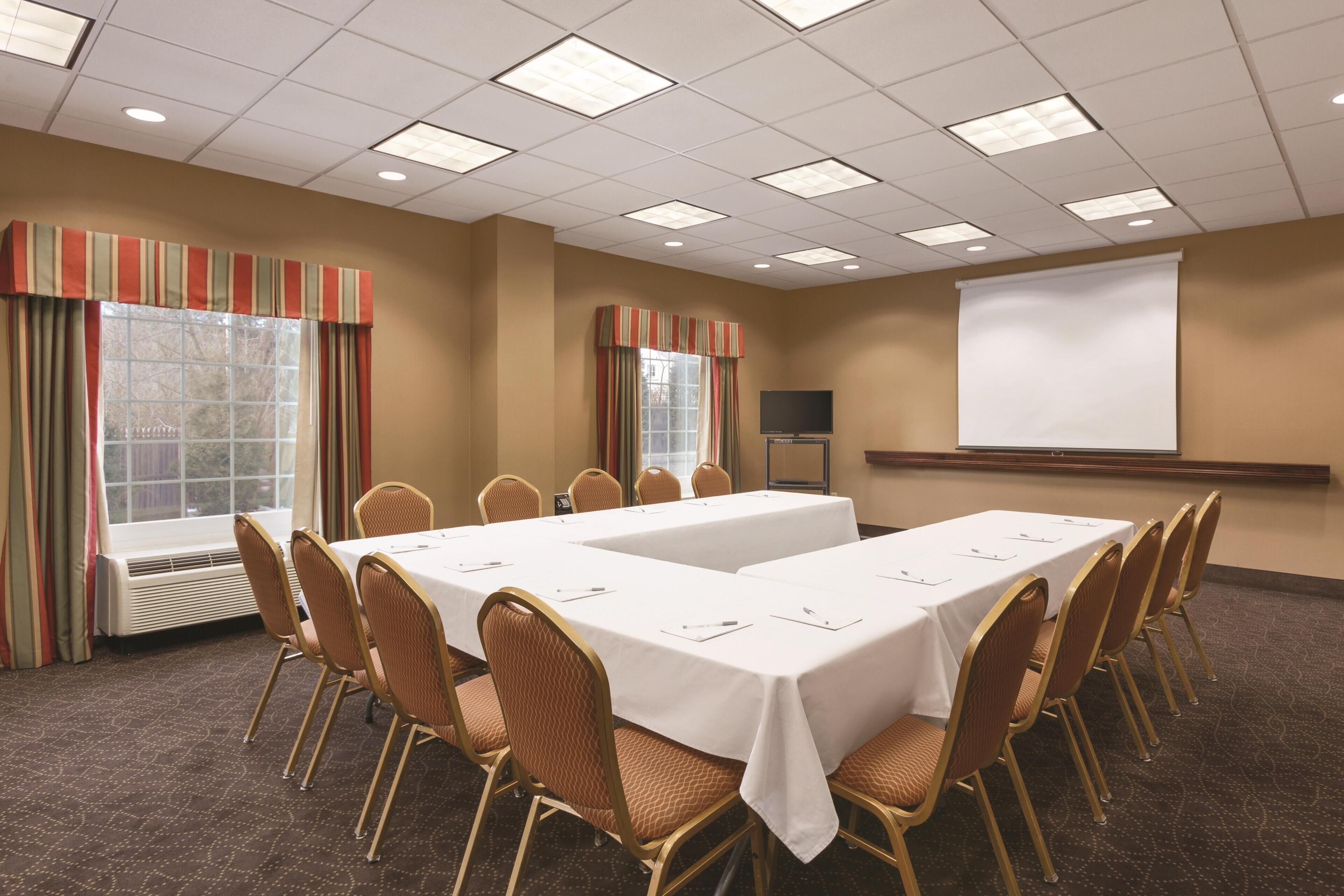 A conference room with a long table, chairs, a projector screen, and windows with curtains.