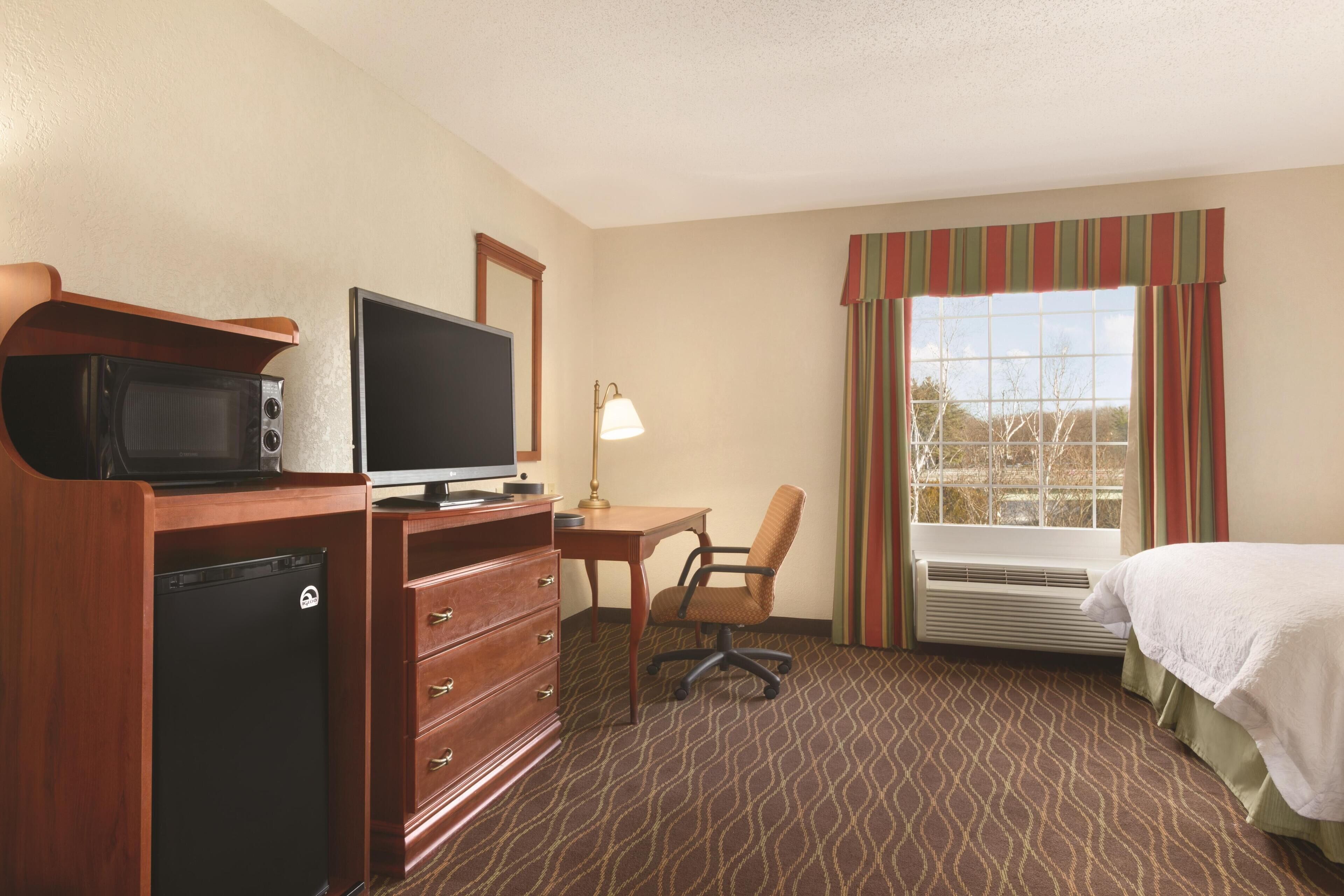 A hotel room with a bed, a desk with a computer, a television, a microwave, a refrigerator, and a window with curtains.