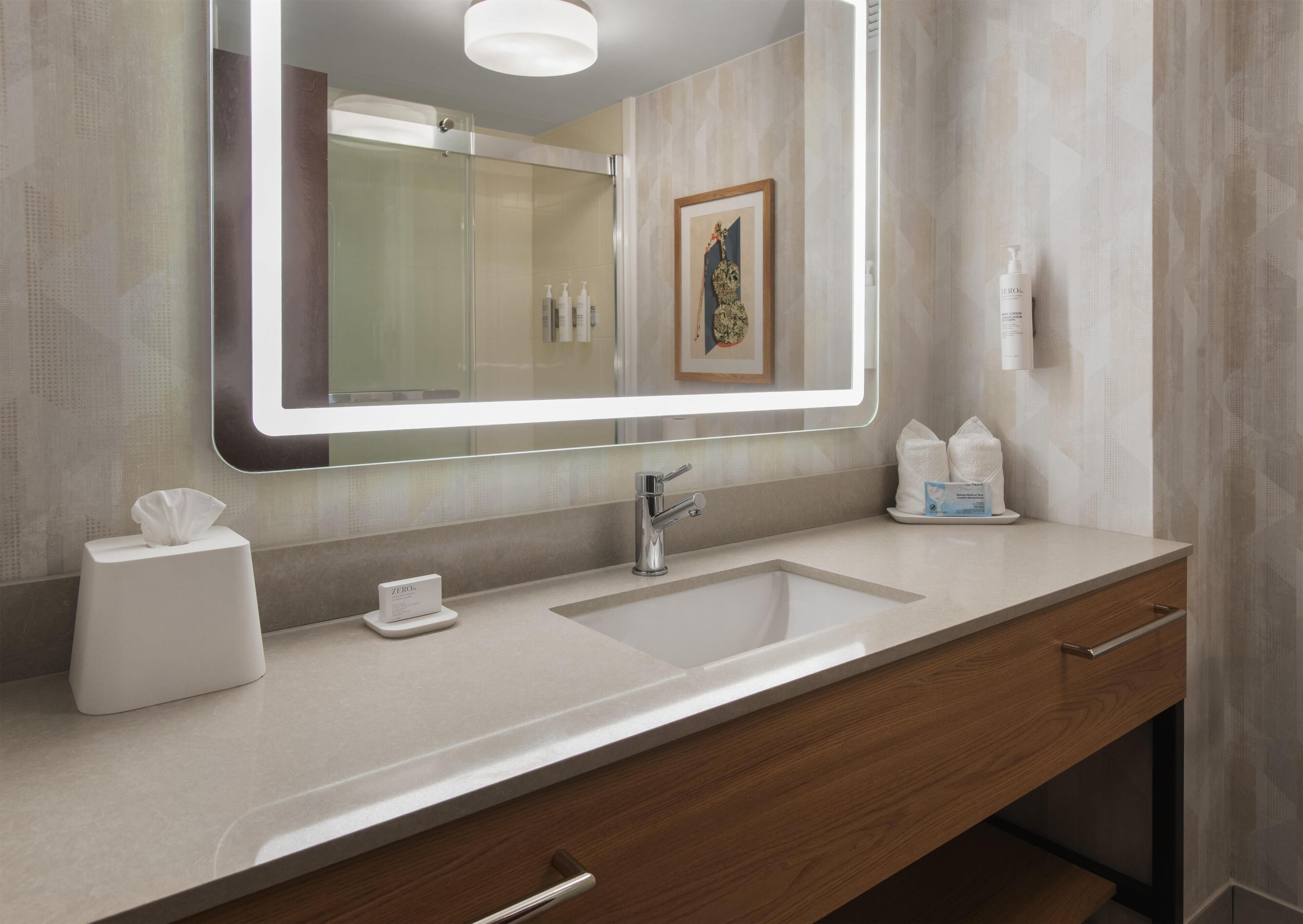 A modern bathroom with a large mirror, two sinks, a towel dispenser, and a framed picture on the wall.