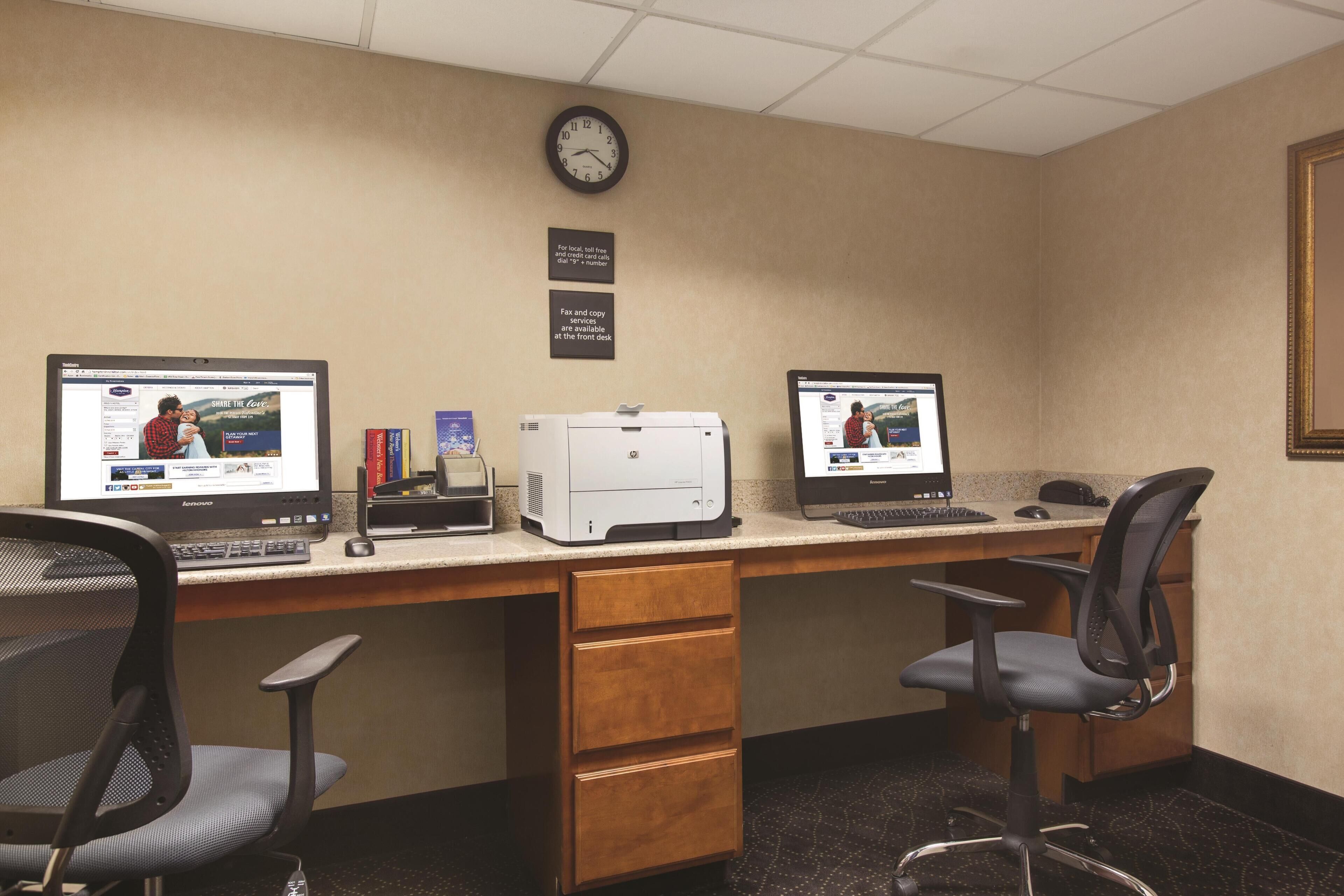 A hotel office with two computer desks, chairs, a printer, and a clock on the wall.