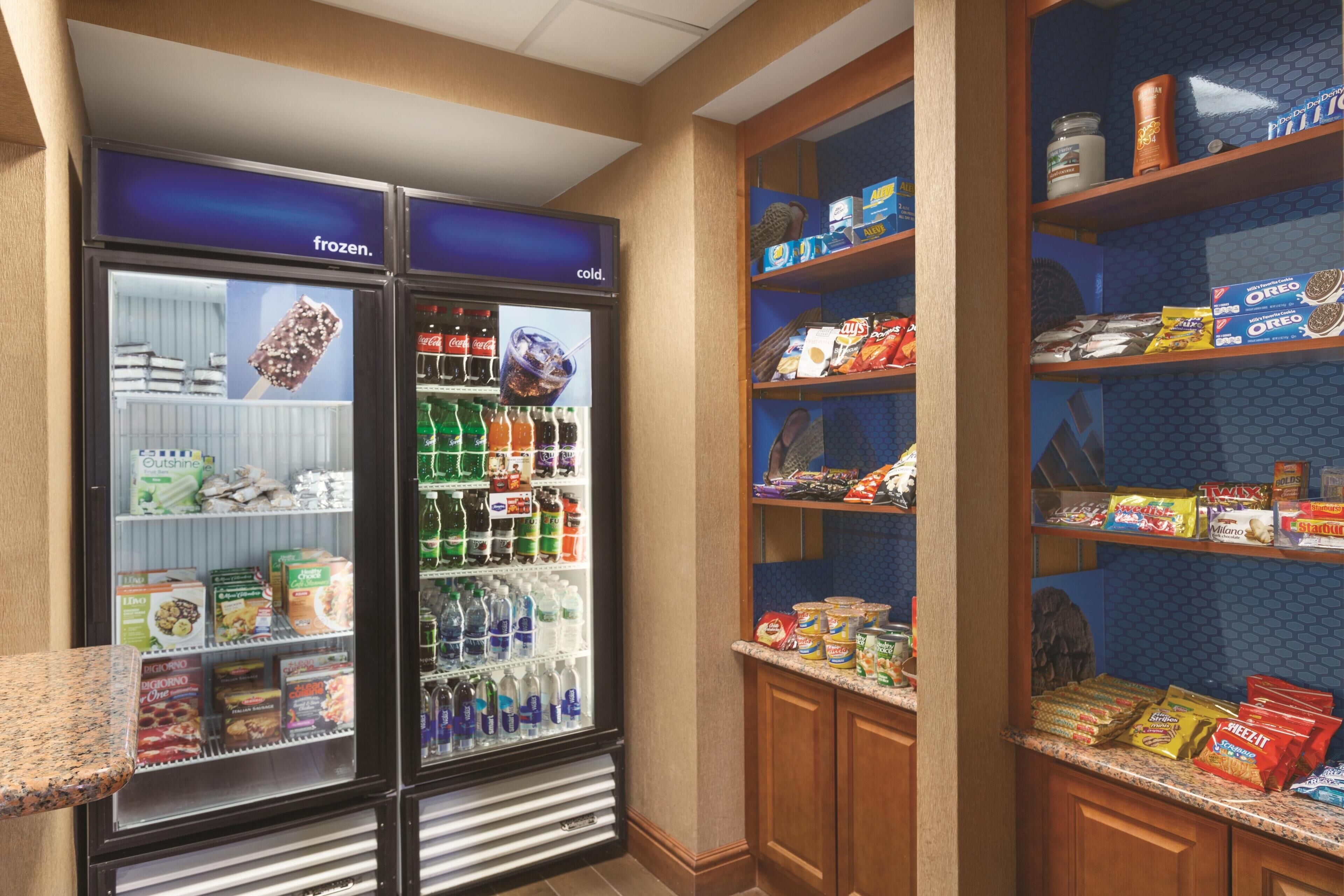 A hotel kitchenette with a refrigerator stocked with snacks, a freezer, and a shelf with packaged food items.