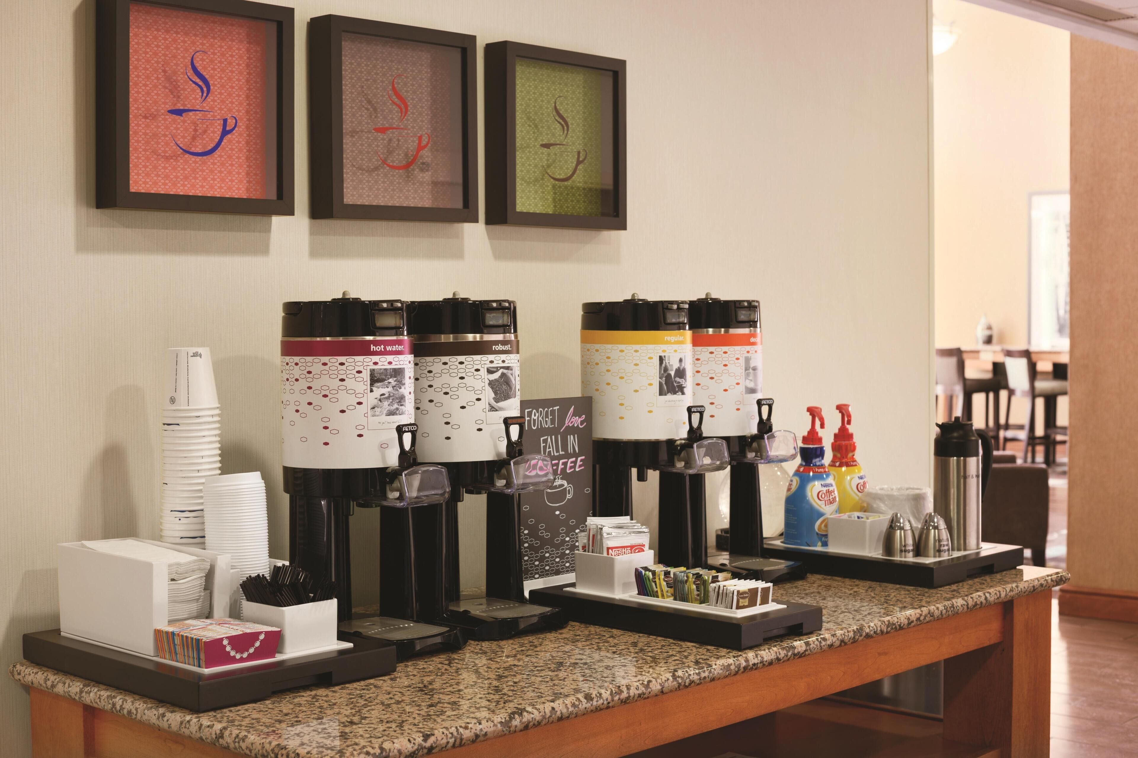A coffee station with multiple dispensers, a stack of white cups, and a sign that says 'Forget love, fall in coffee.'