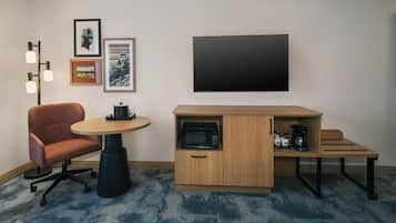 Quarto de hotel compacto com uma mesa redonda, uma cadeira estofada, uma kitchenette com micro-ondas, máquina de café e um pequeno armário.