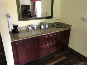 Un baño con encimera de granito, un lavamanos, un espejo y un televisor sobre el espejo.