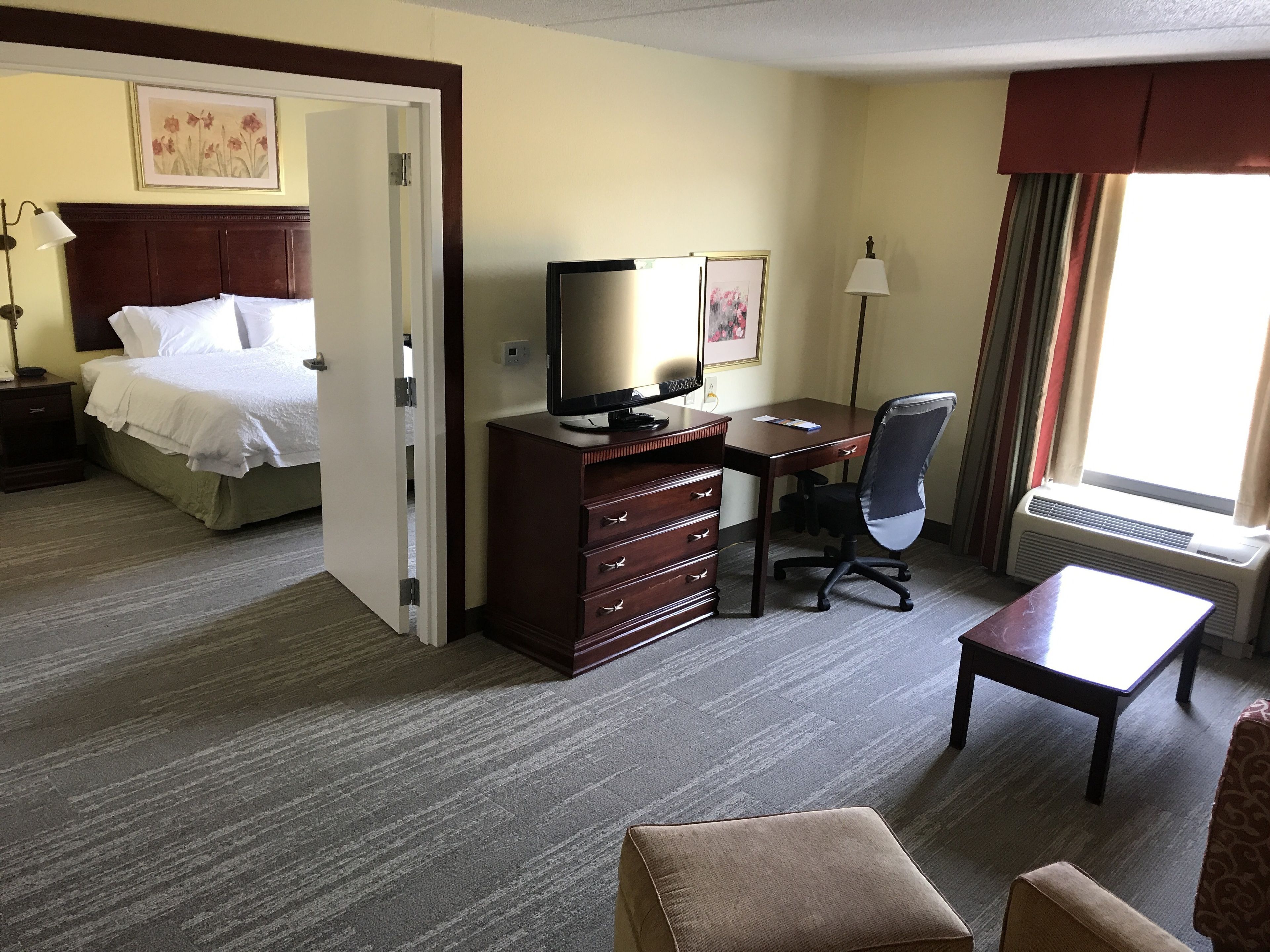 Quarto de hotel com cama, secretária com computador, cadeira, mesinha e janela com cortinas.