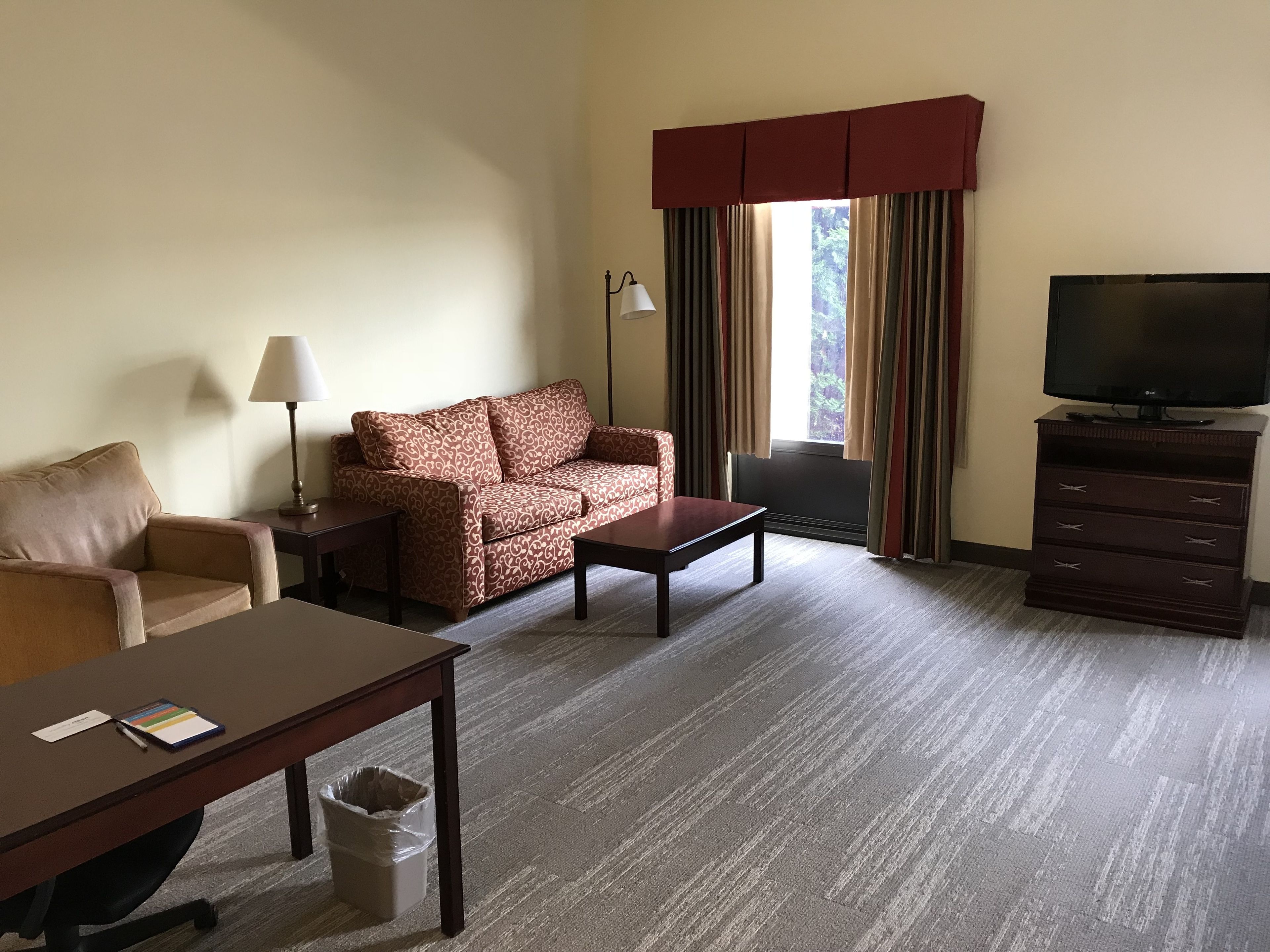 A hotel room with a sofa, a small table, a TV, a lamp, and a window with curtains.