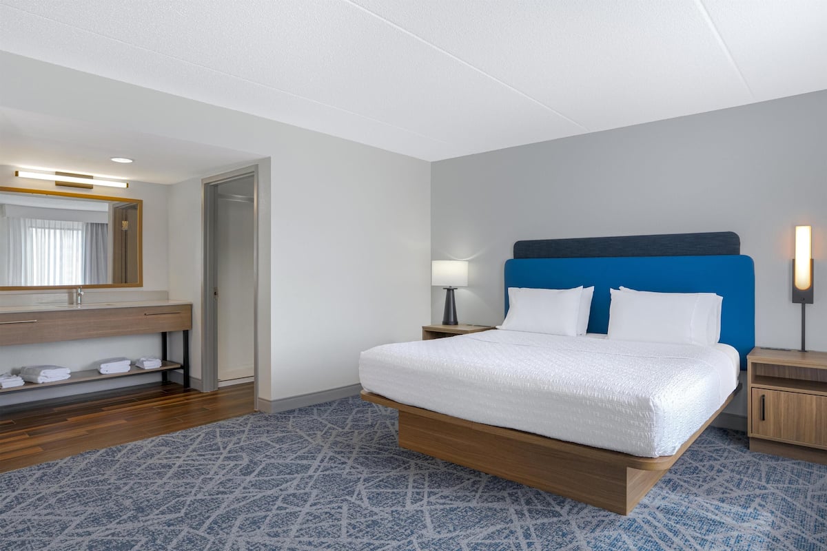 A hotel room with a bed, a nightstand, a lamp, a mirror, a wooden dresser, and a patterned rug.