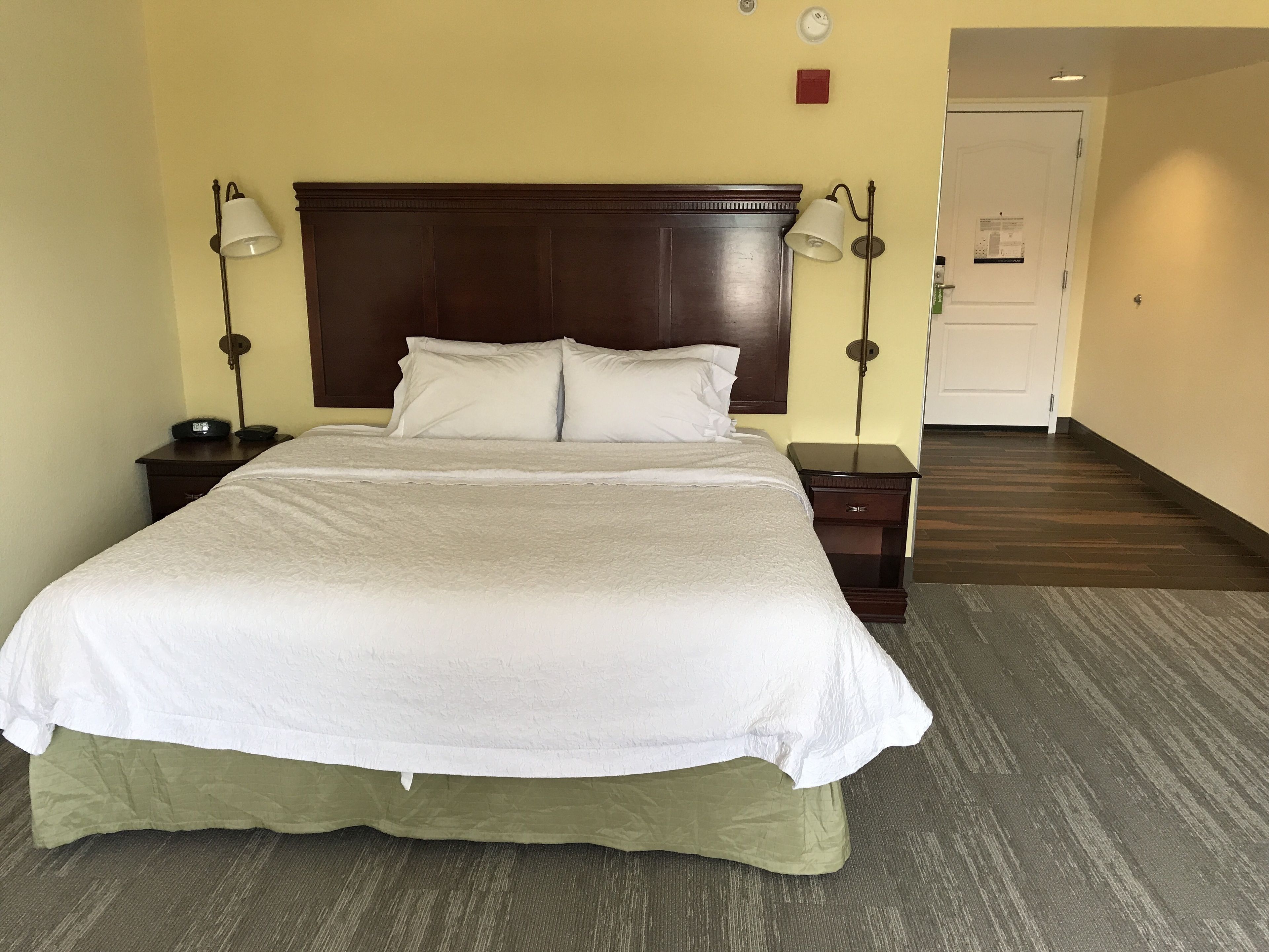 A hotel room with a large bed, two bedside tables, a wooden headboard, and a carpeted floor.