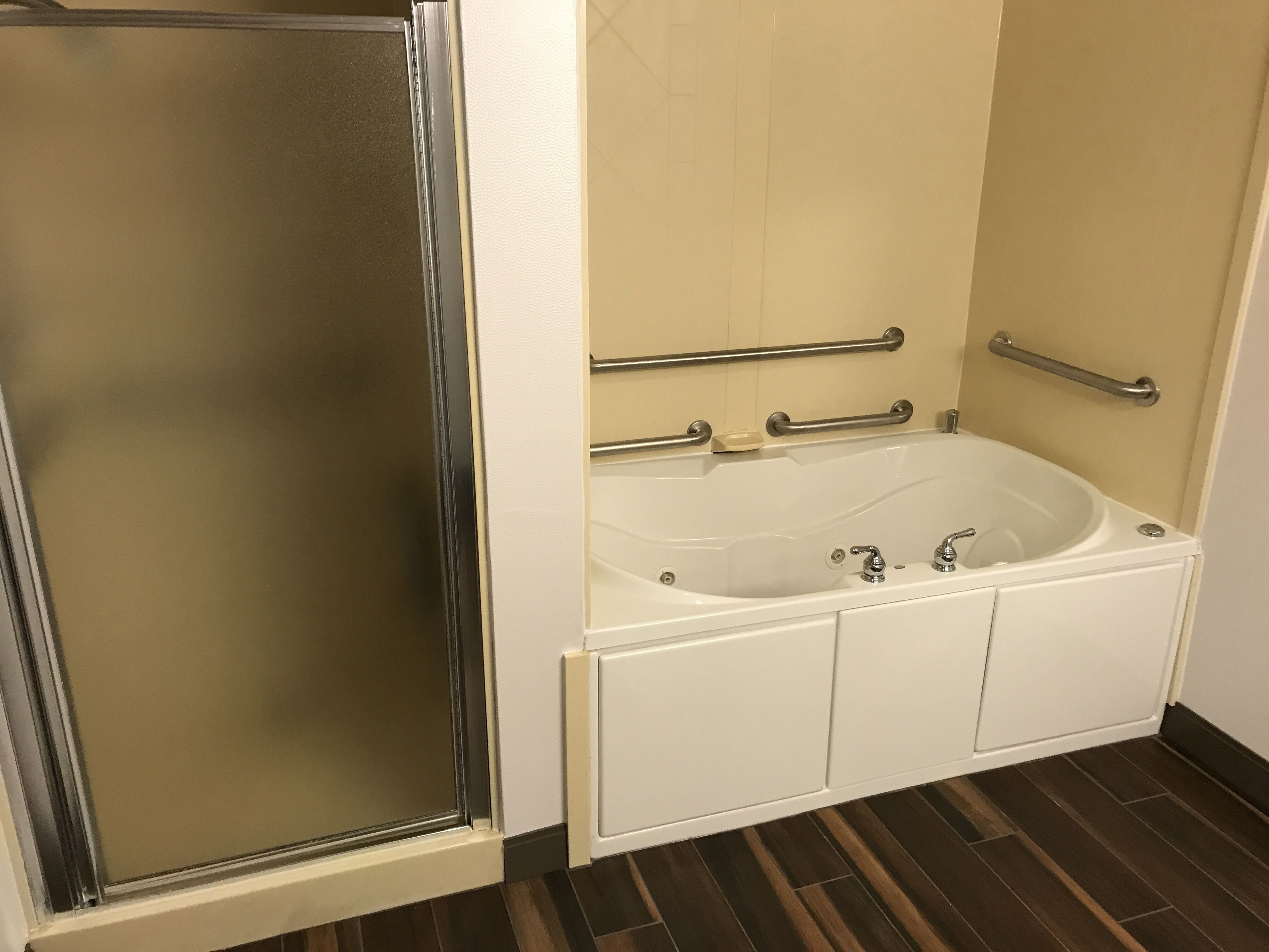A bathroom with a glass shower enclosure, a white bathtub with grab bars, and a dark wood floor.