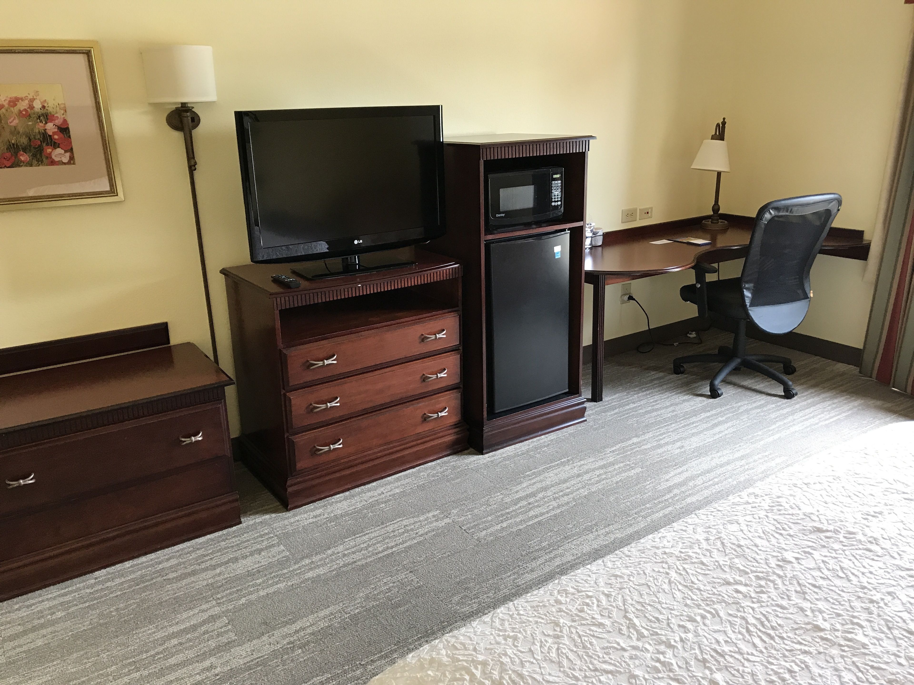 A hotel room with a wooden dresser, a flat-screen TV, a mini-fridge, a desk with a lamp, and a black office chair.
