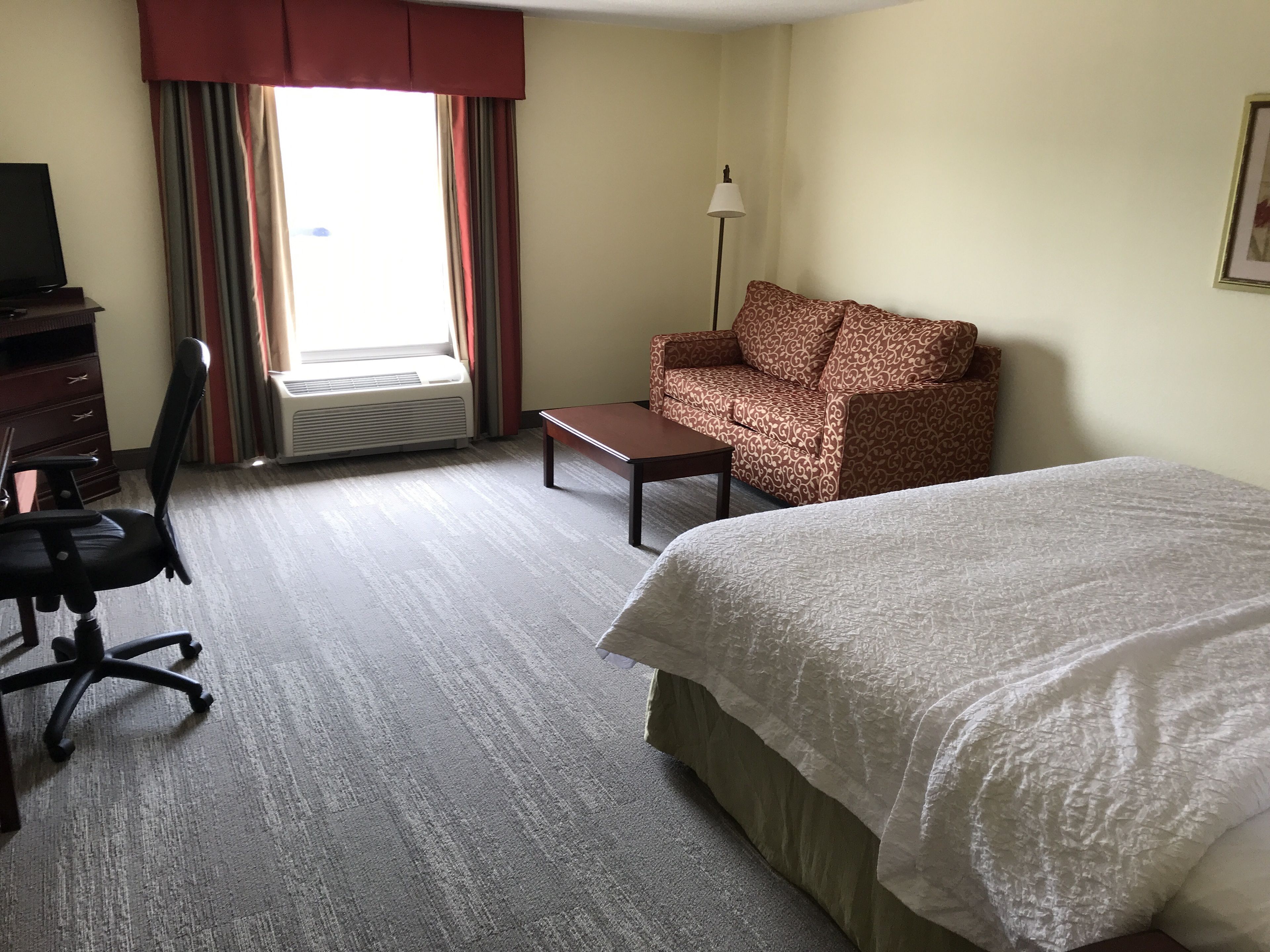 Une chambre d’hôtel comprenant un lit, un bureau avec une chaise, un canapé, un lampadaire et une fenêtre avec des rideaux.
