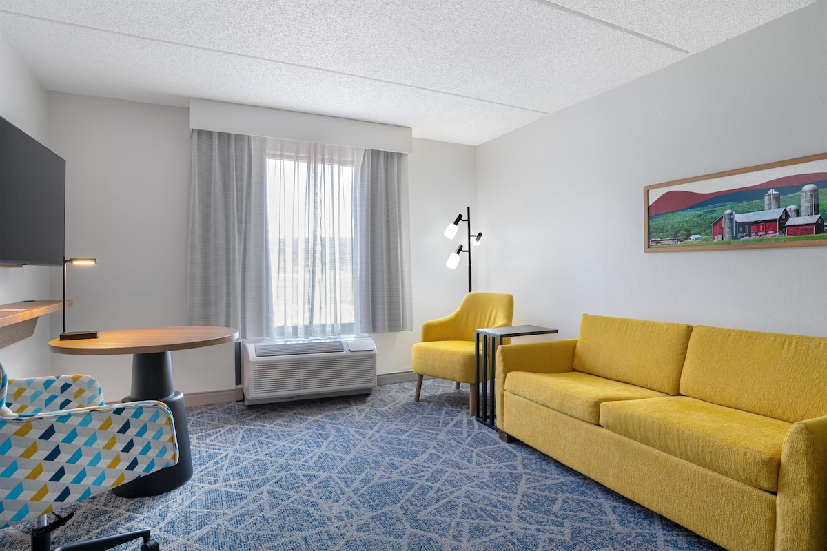 A hotel room with a yellow sofa, a yellow armchair, a small round table, a TV on a stand, a large window with curtains, and a painting on the wall.