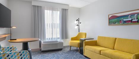 A hotel room with a yellow sofa, a yellow armchair, a small round table, a TV on a stand, a large window with curtains, and a painting on the wall.